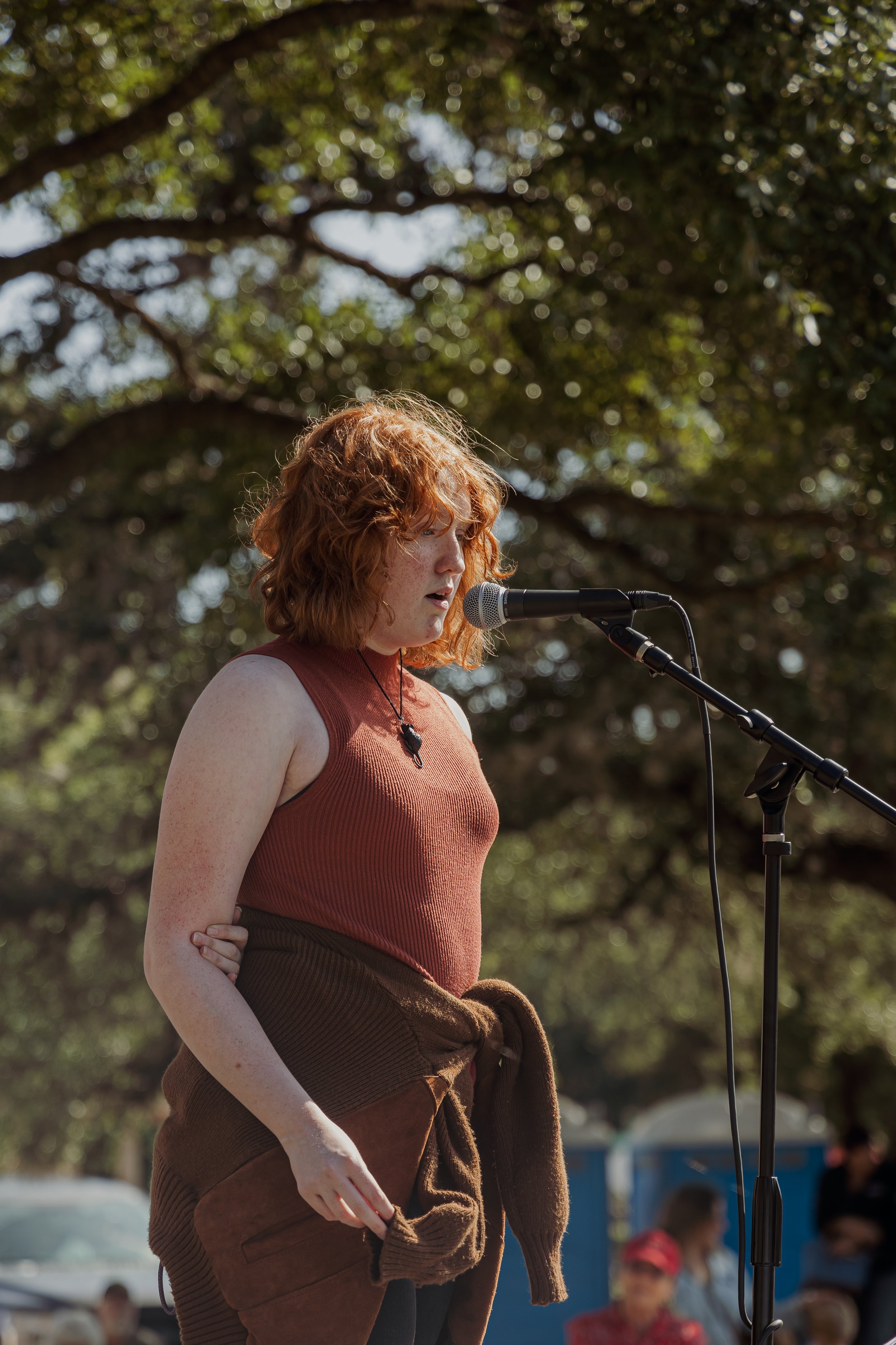 Red-haired woman speaking into a microphone outdoors with trees in the background.