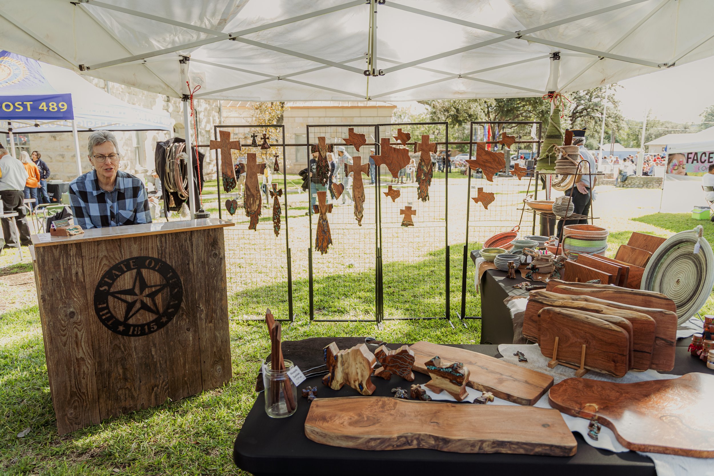 An outdoor craft vendor booth with wooden art, jewelry, and Texas-shaped decorations, under a canopy at a fair or market.