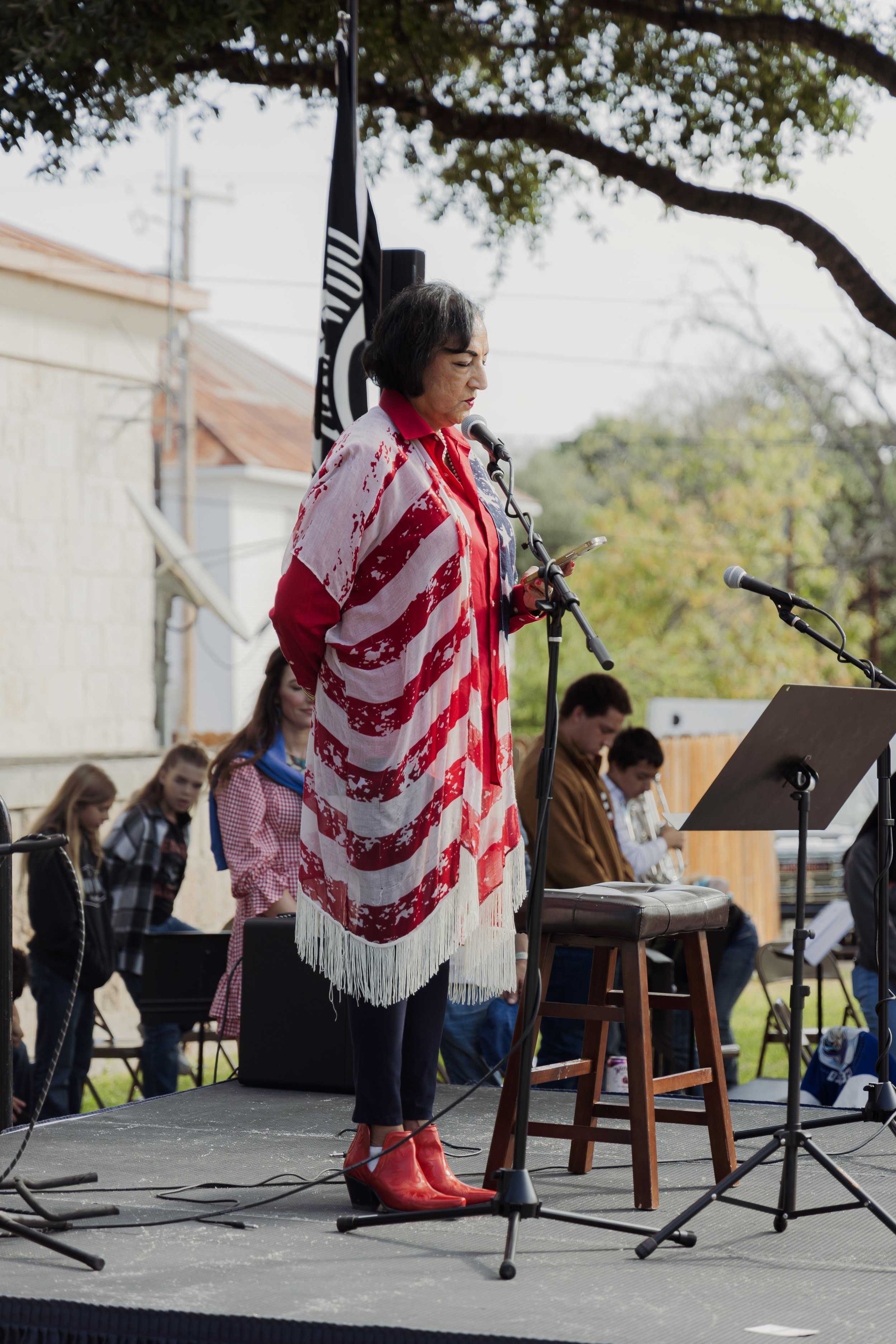 A woman wearing red boots and a red, white, and blue striped shawl speaking into a microphone at an outdoor event, with a group of people seated behind her.