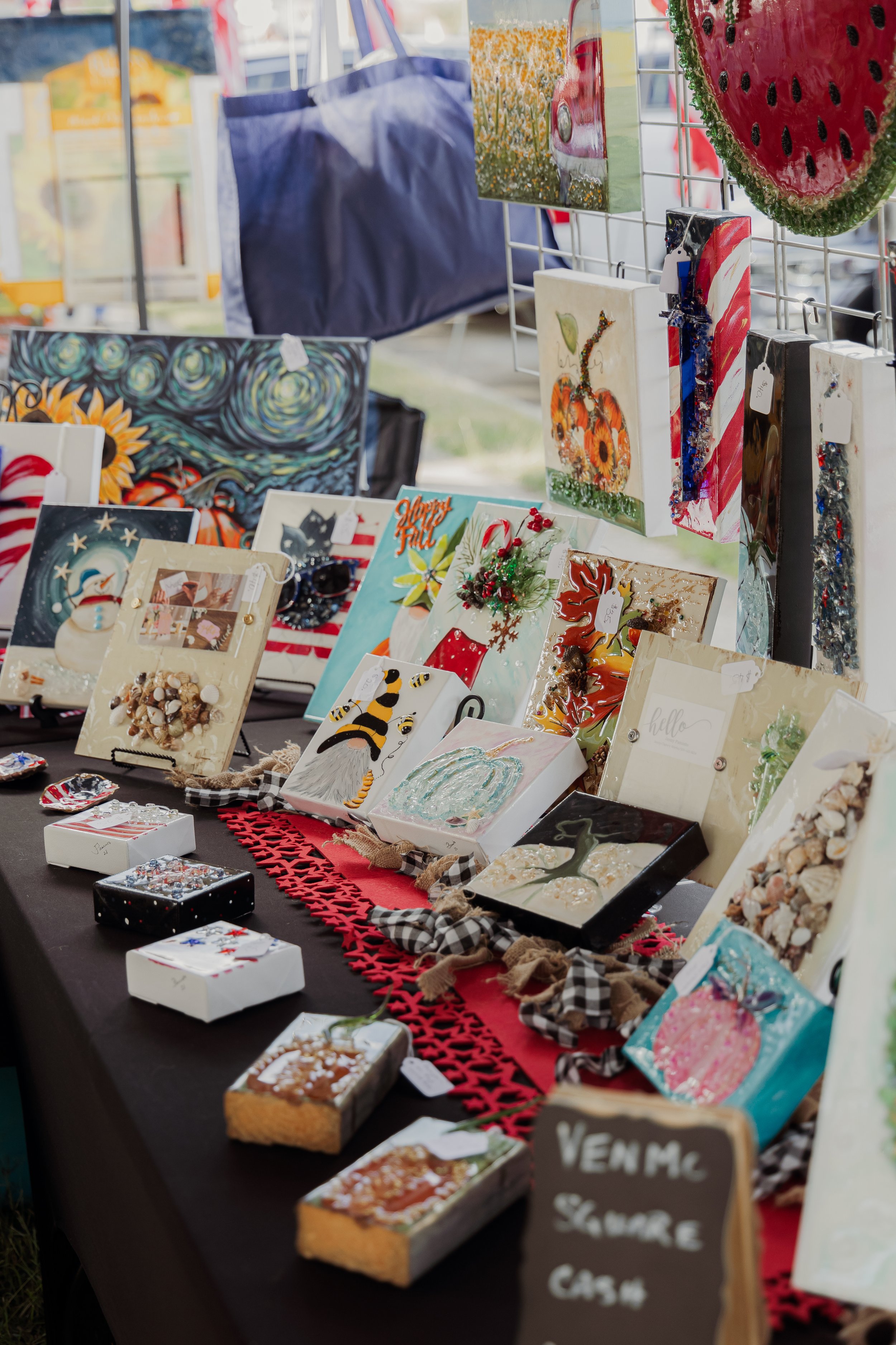 A display of handmade holiday-themed paintings and crafts on a table at an outdoor market, with some paintings depicting snowmen, pumpkins, and abstract art, and small decorated boxes and baked goods in front.