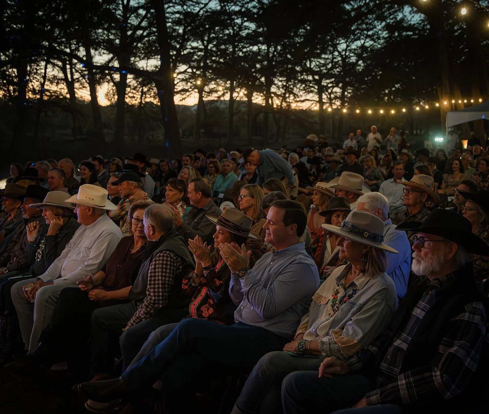 An outdoor evening concert or event with a large audience seated under string lights among trees, sunset in the background.