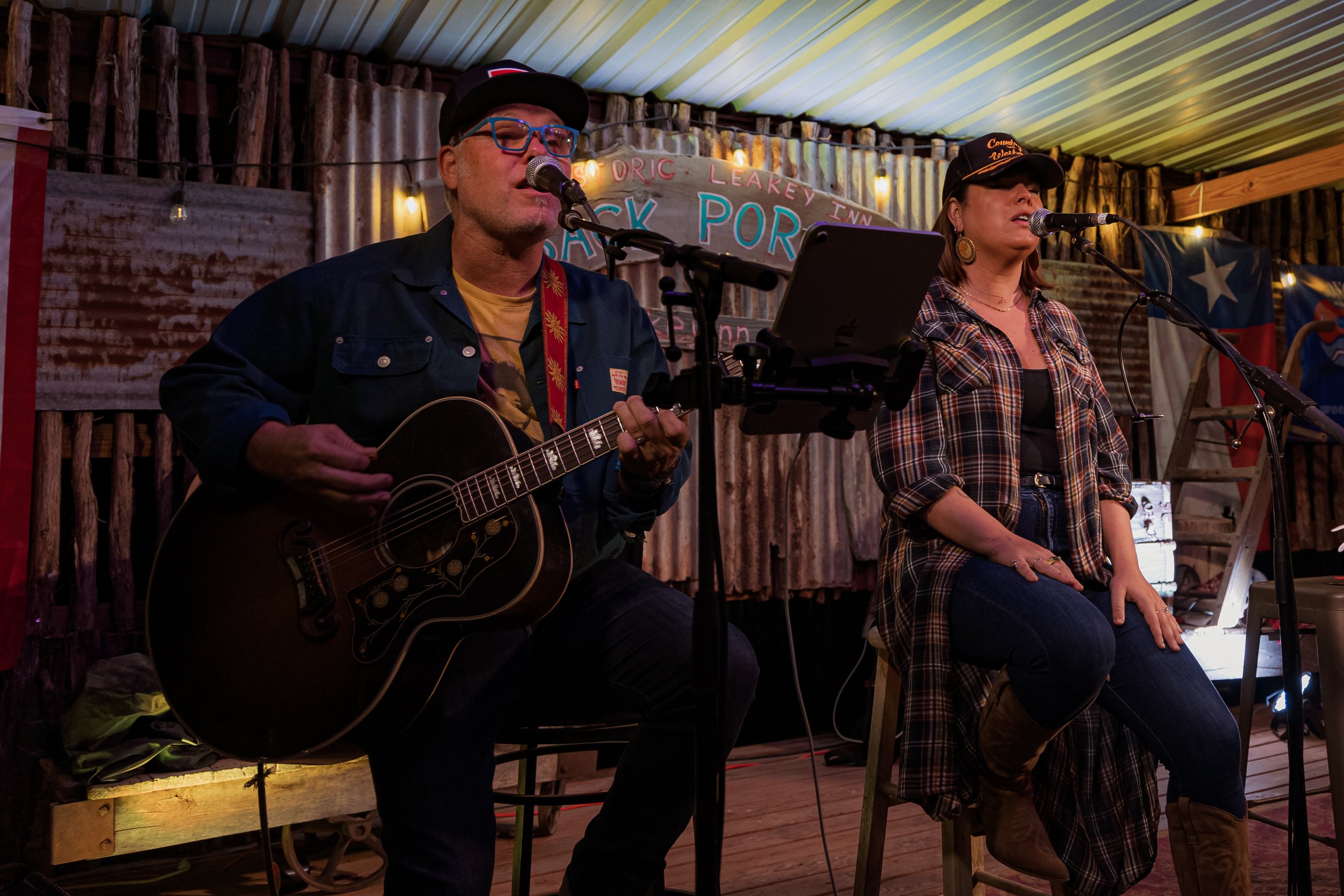 Two musicians performing on a rustic stage. The man on the left is playing an acoustic guitar and singing into a microphone. The woman on the right is sitting on a stool, also singing into a microphone. The stage is decorated with string lights, corr