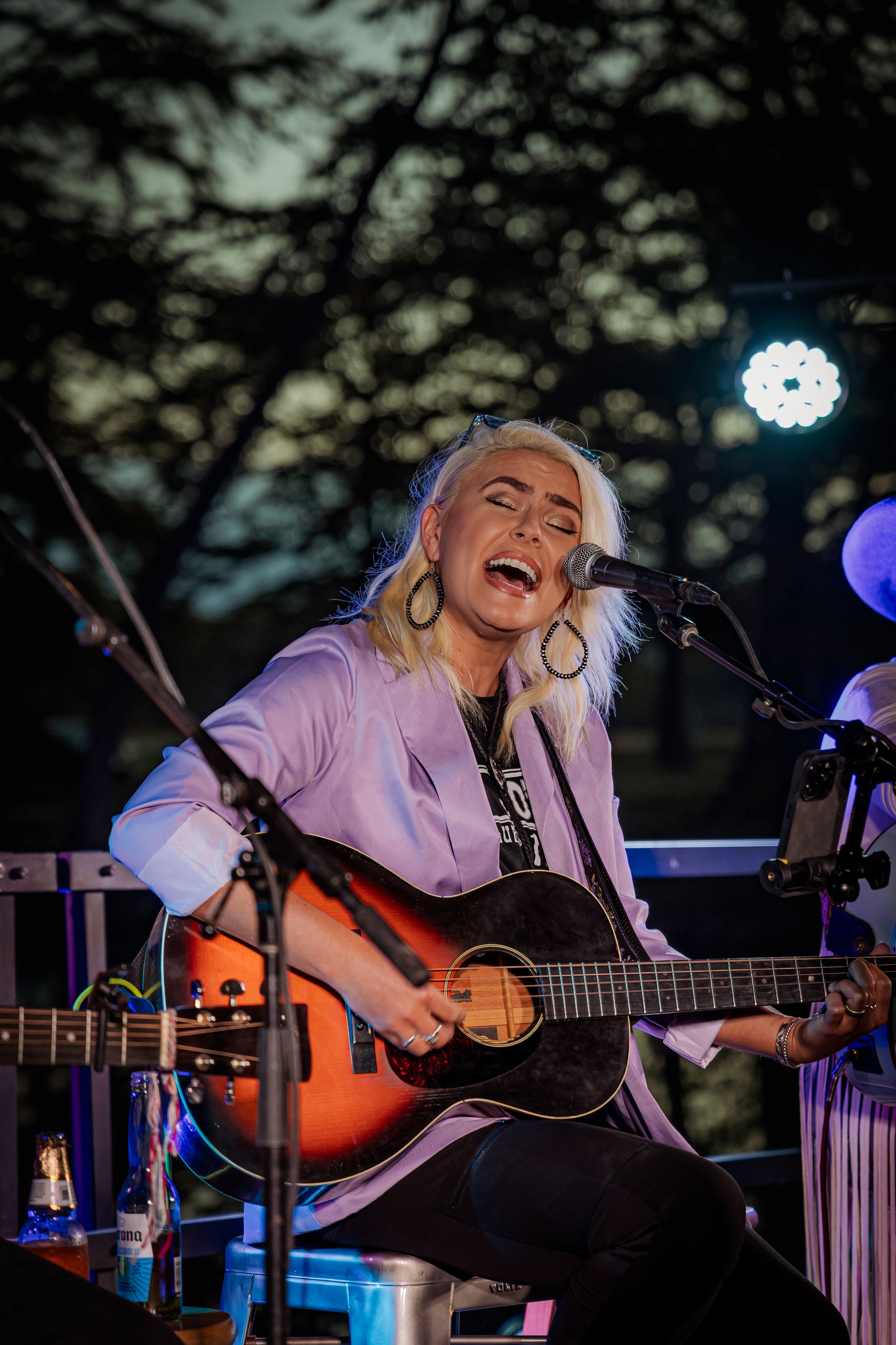 A woman singing and playing an acoustic guitar during a live outdoor performance at night, with dark trees and stage lights in the background.