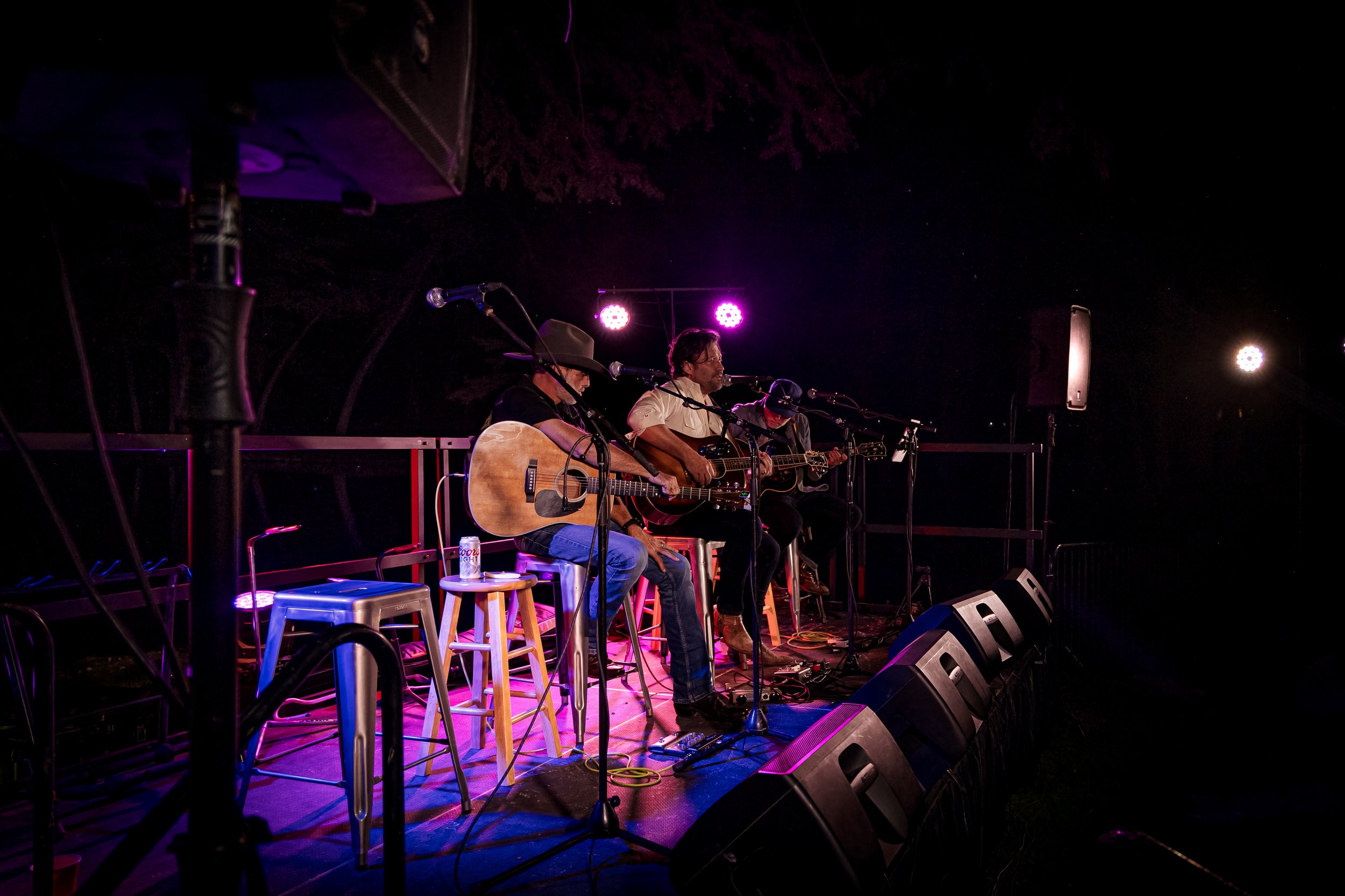 Three musicians performing on a dimly lit outdoor stage at night, with guitars and microphones, surrounded by purple and white lights.