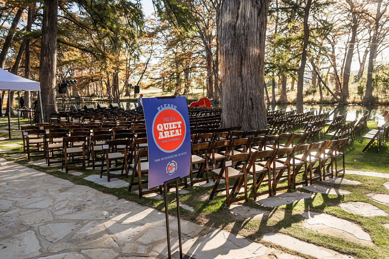 Empty outdoor seating area with numerous wooden chairs facing a stage near a river, surrounded by large trees. A sign in the foreground reads, 'Please, this is a quiet area during performances,' indicating the space is for a performance or event.
