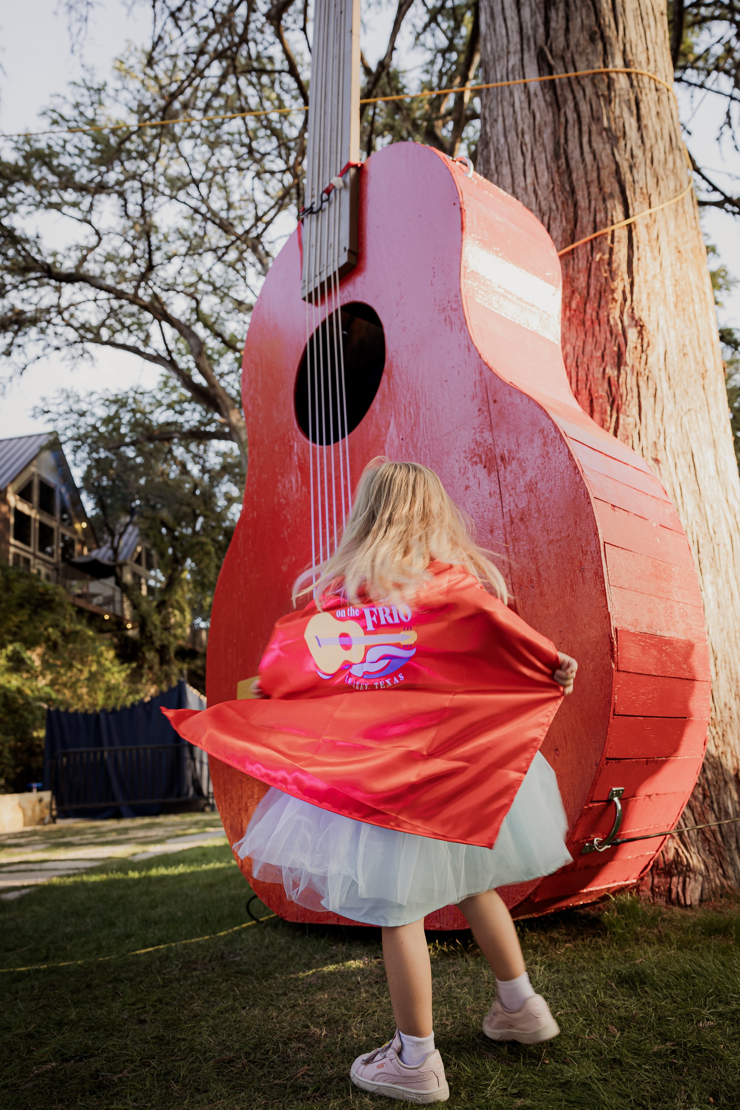 A young girl wearing a red jacket with a guitar logo on the back, a blue skirt, and sneakers, tugging on her jacket, standing in front of a large, red, guitar-shaped structure attached to a tree outdoors.