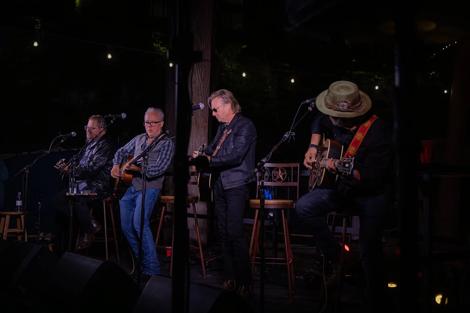 Four musicians performing on stage outdoors at night, each playing an acoustic guitar, with three of them standing and one seated, surrounded by dark trees with string lights overhead.