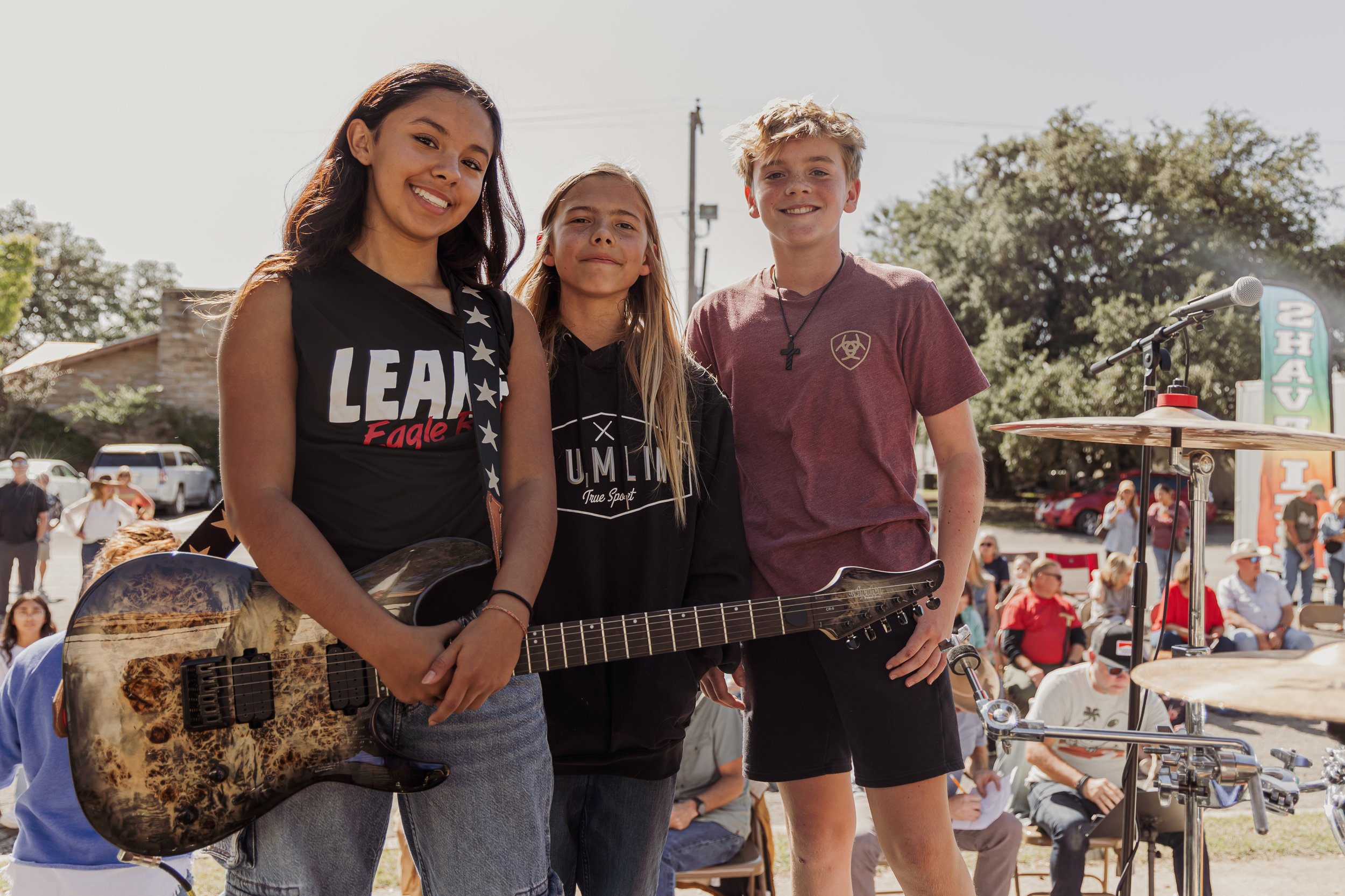Three young people standing together outdoors at a music event, with a crowd and trees in the background.