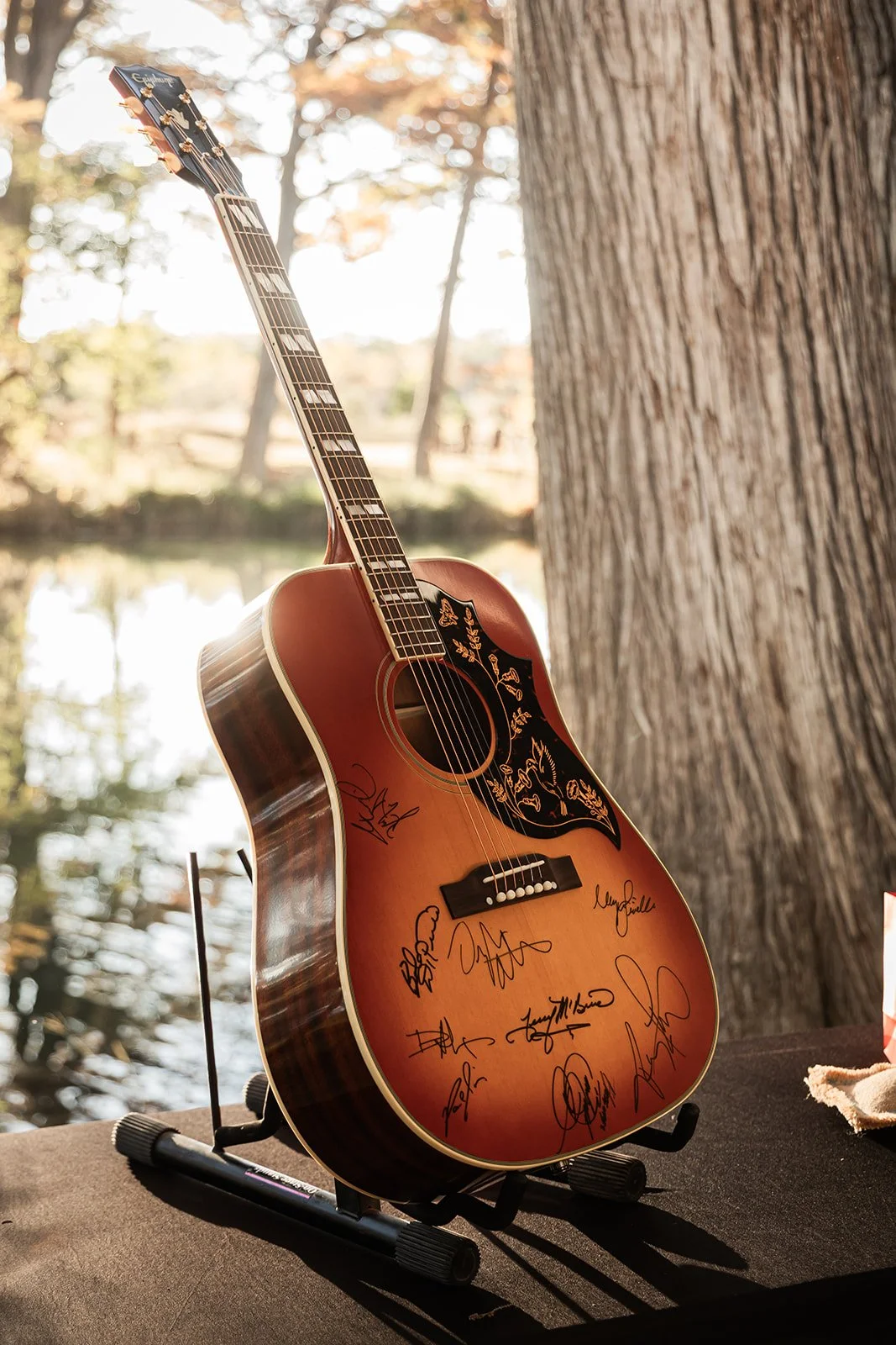 An autographed acoustic guitar resting on a black stand outdoors near a large tree, with a body of water and trees in the background.