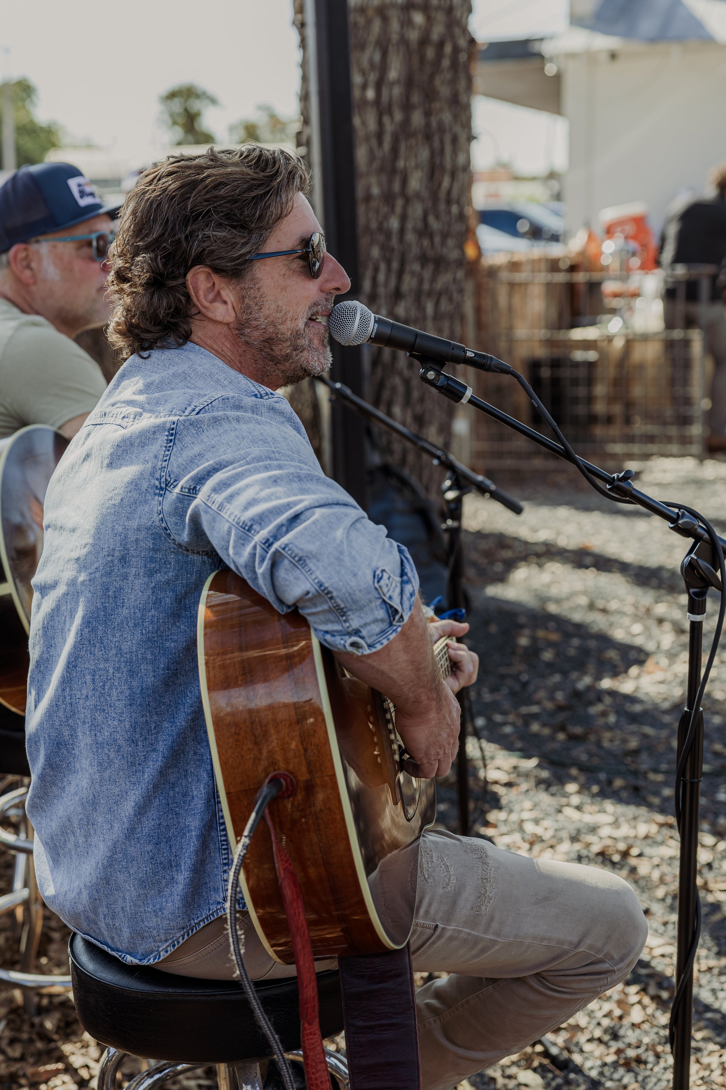 Man with sunglasses playing an acoustic guitar and singing into a microphone outdoors at a sunny event.