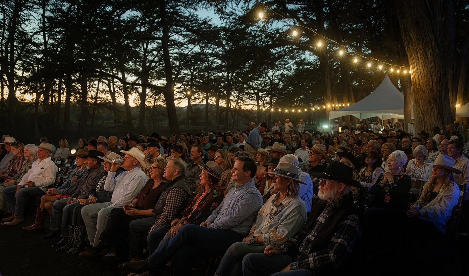 An outdoor gathering at dusk with many people seated and watching an event, illuminated by string lights, surrounded by trees and tents.