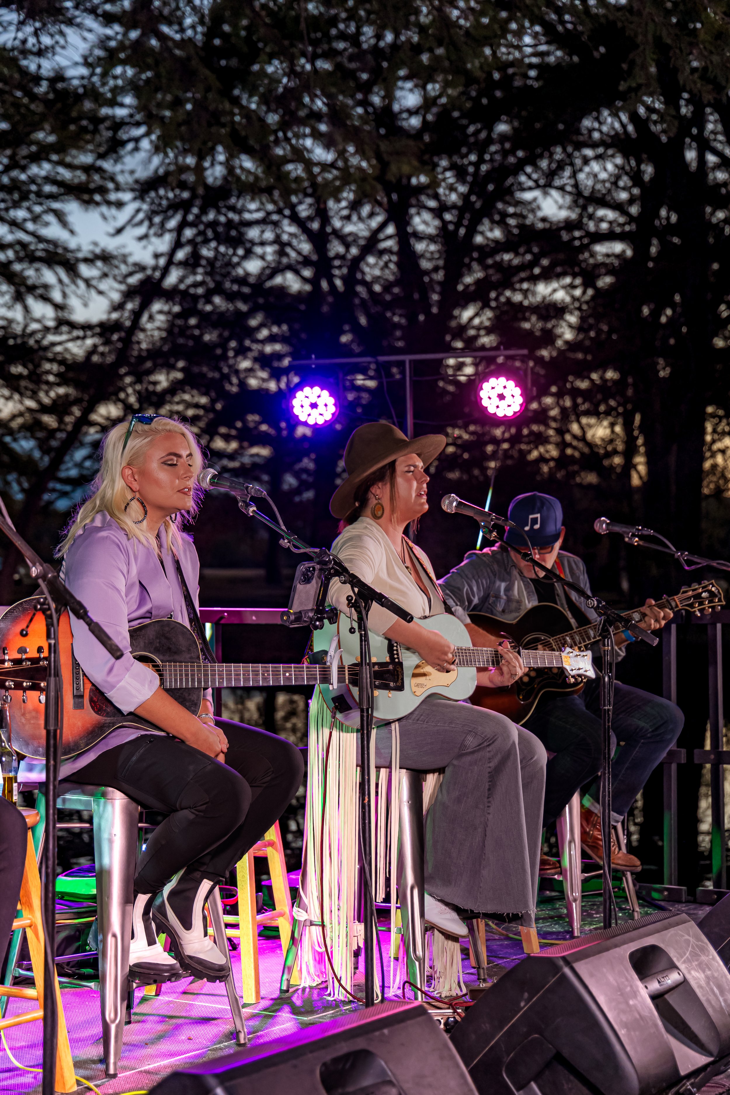 Three musicians performing on an outdoor stage at dusk, two women and one man, playing guitars and singing into microphones, with purple stage lights and trees in the background.