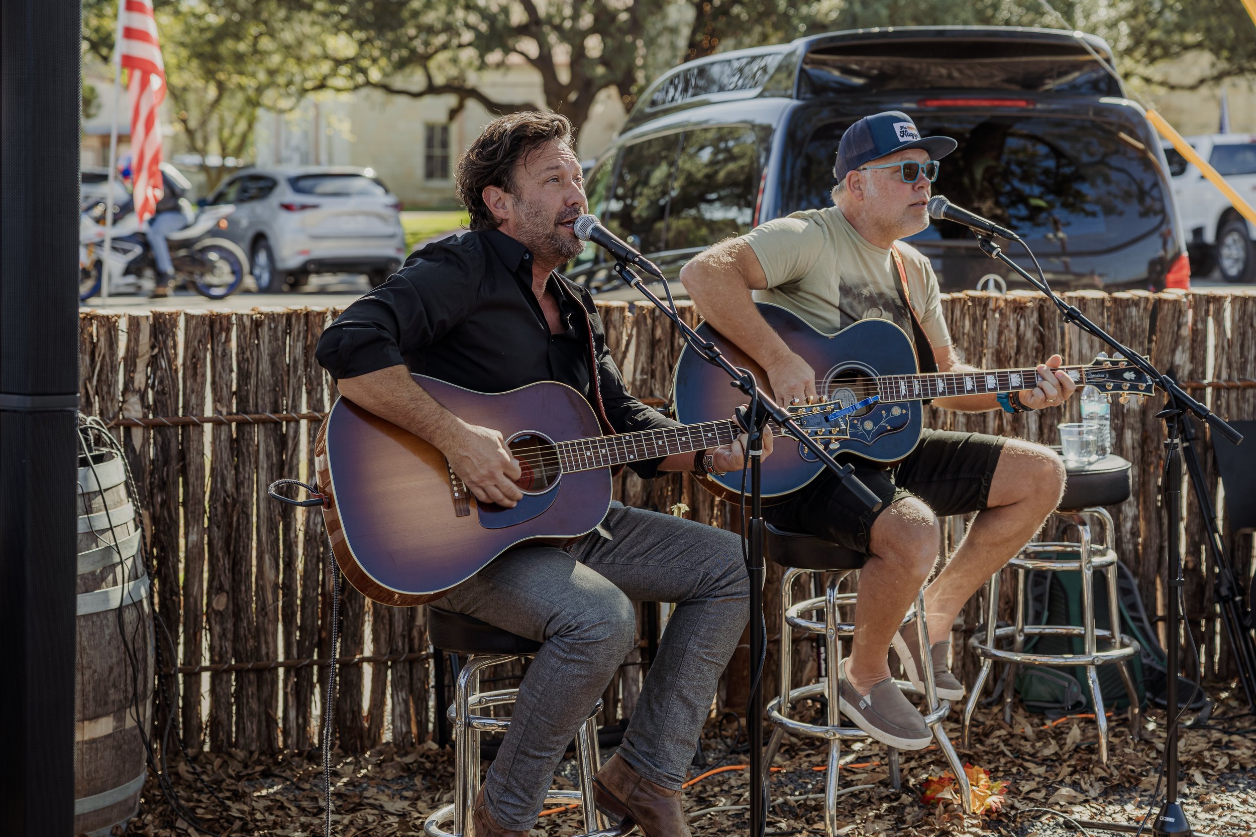 Two men are playing acoustic guitars and singing into microphones outdoors during the day. They are seated on stools with a rustic fence behind them, and parked cars are visible in the background.
