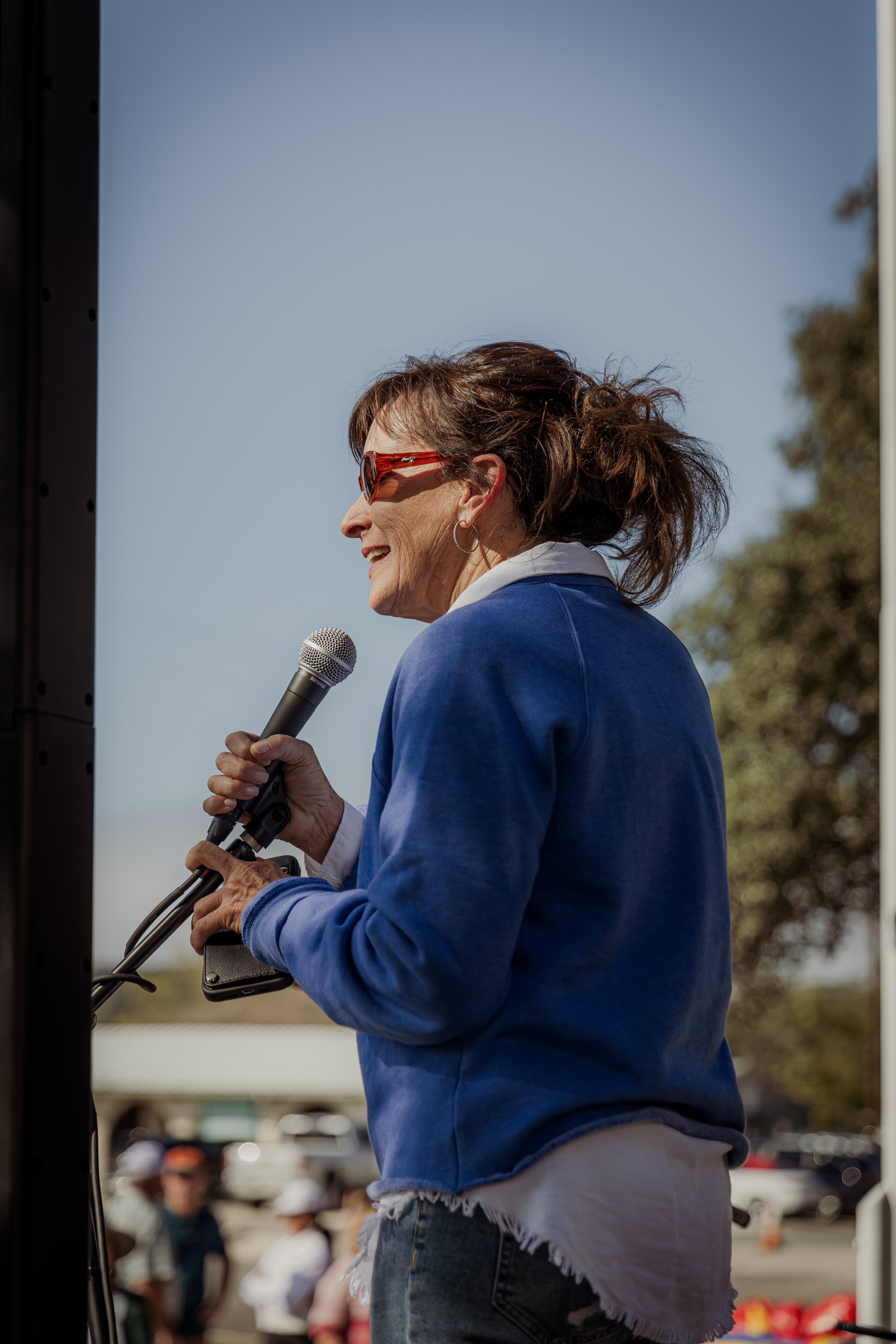 A woman wearing sunglasses and a blue jacket is speaking into a microphone outdoors during the daytime, with a group of people and trees in the background.