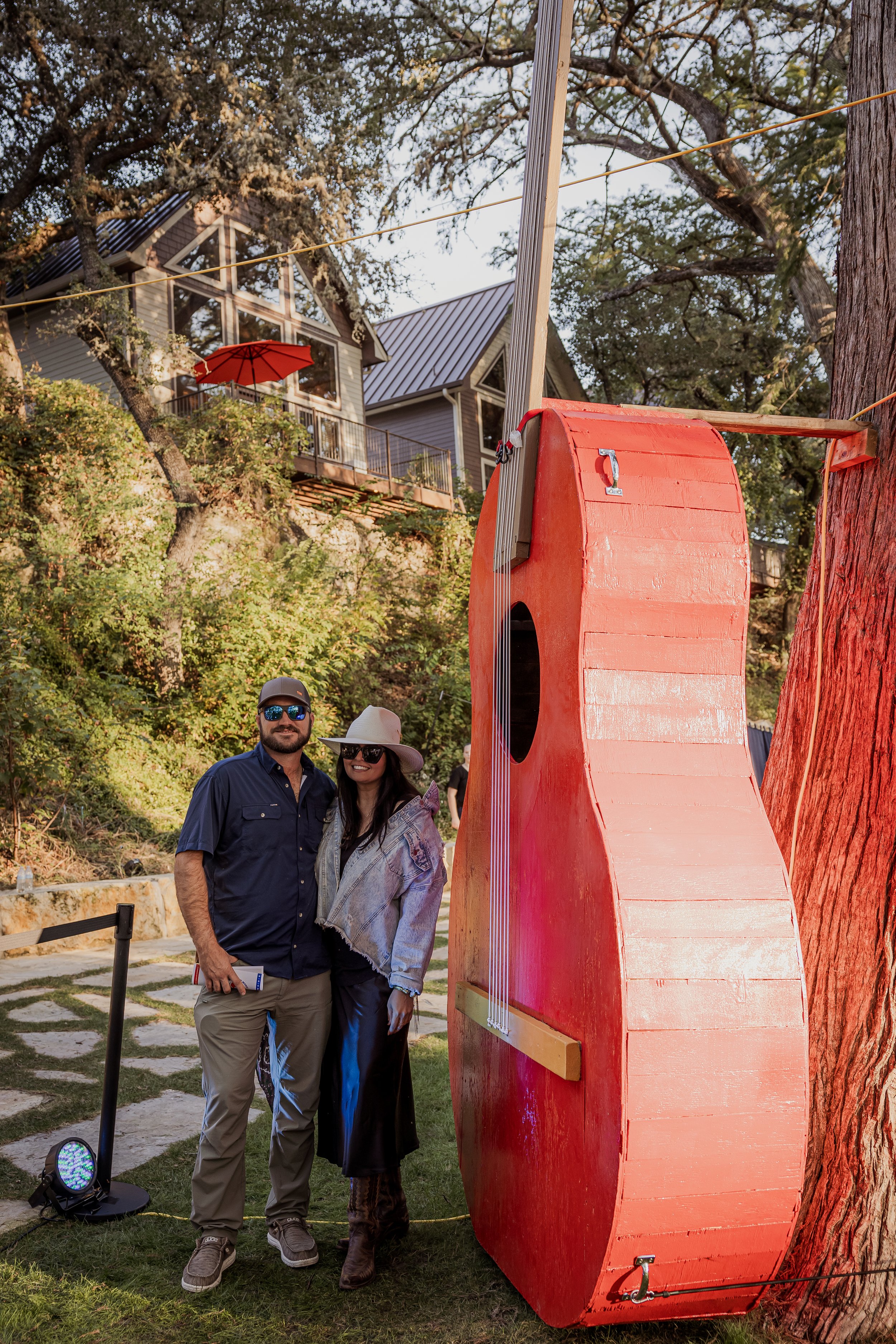 A man and woman stand together outdoors near a large, red, wall-mounted guitar-shaped structure, with houses and trees in the background.