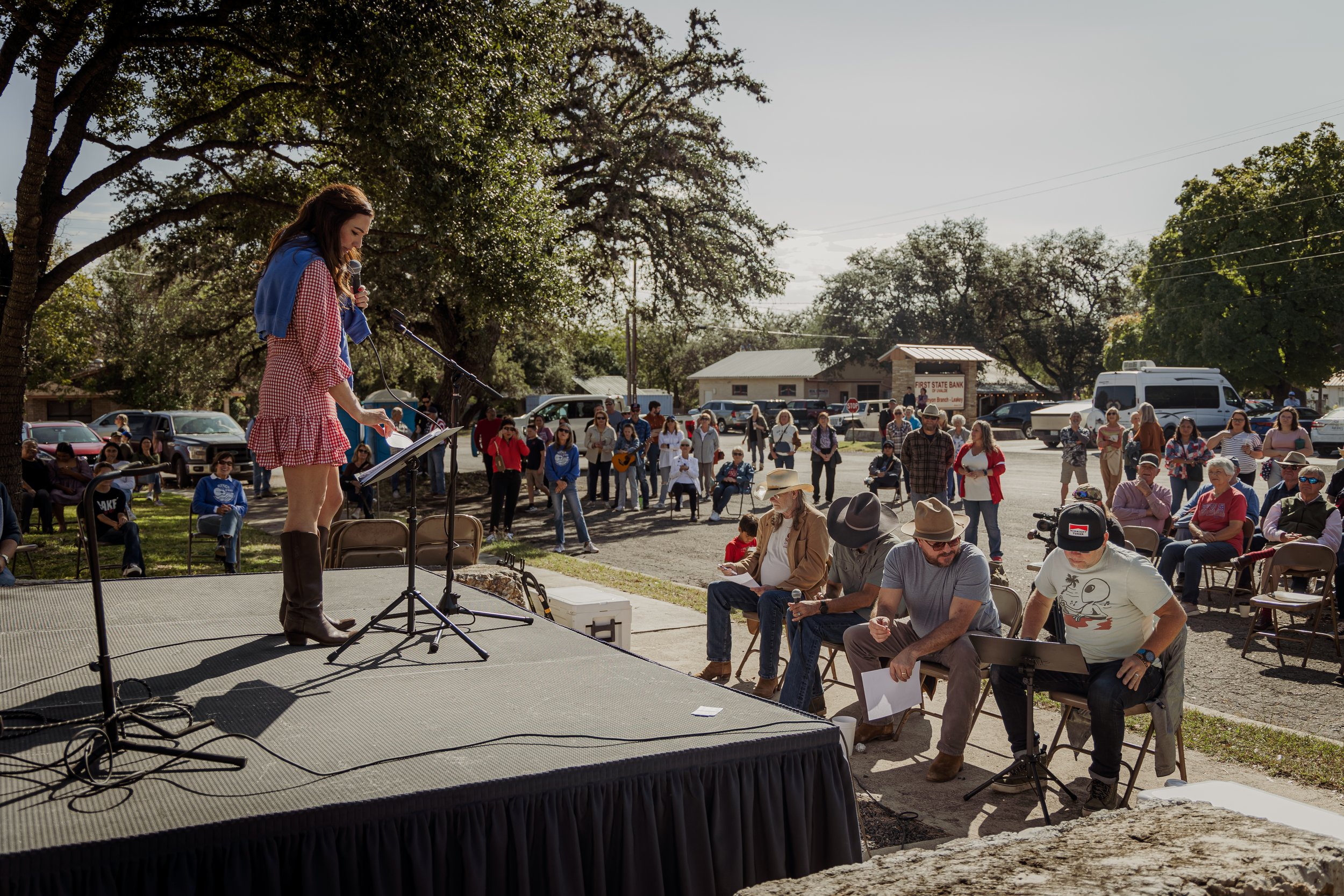 A woman in a red and white checkered dress and brown boots speaks into a microphone on stage, while a large group of people watch outdoors on a sunny day, gathered around and sitting on chairs in front of the stage.