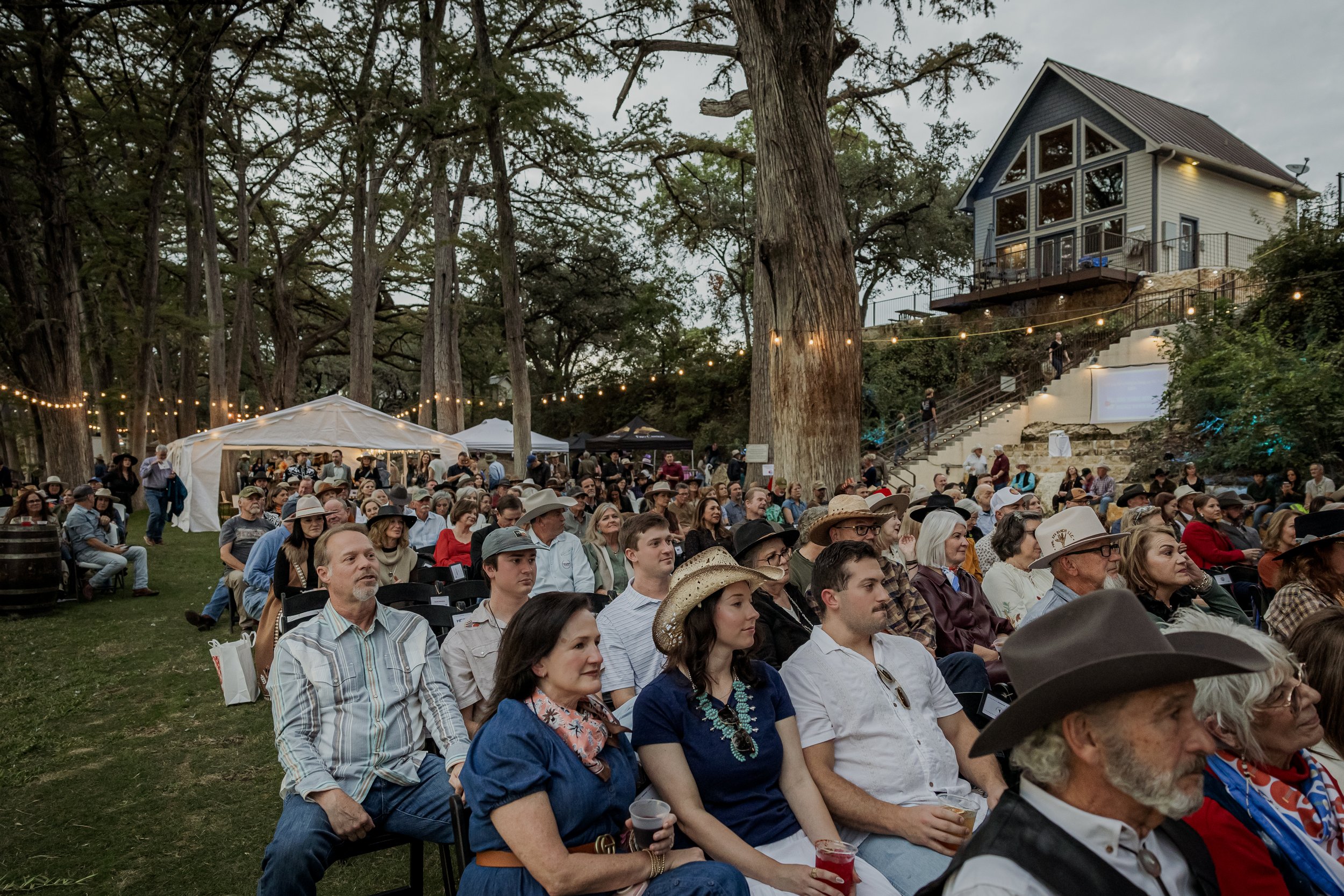 An outdoor gathering of people seated on chairs in a wooded area, attending a concert or event during the evening with string lights overhead and a house on a hill in the background.