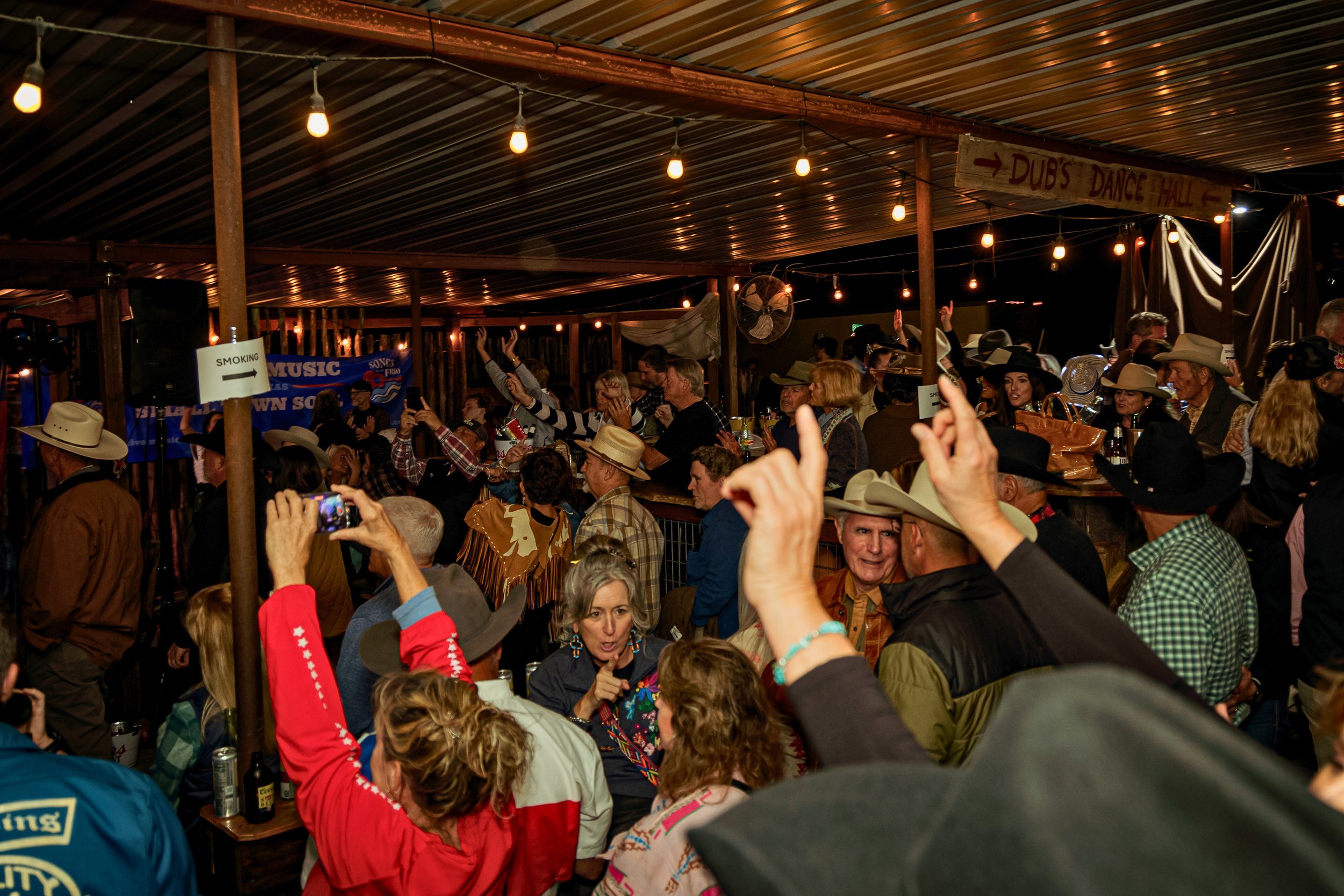 Crowd of people at a cowboy-themed country dance party, many wearing cowboy hats, inside a dimly lit venue decorated with string lights and a wooden sign pointing to 'Dub's Dance Hall'.