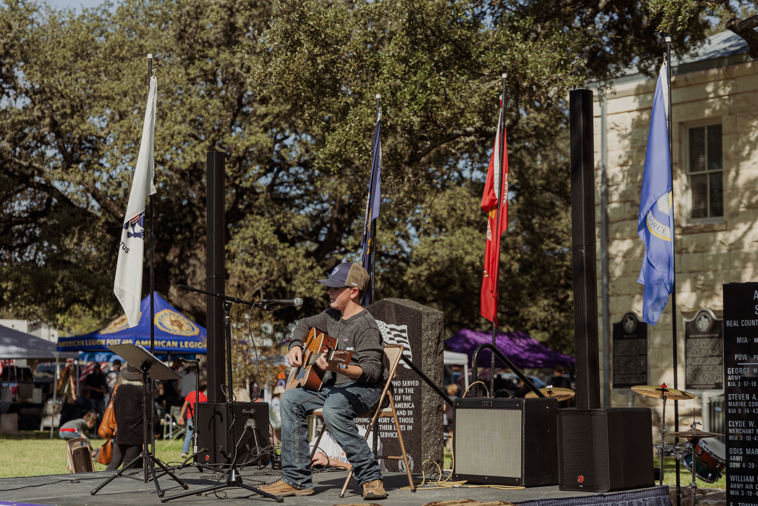 A young boy playing an acoustic guitar and singing on a stage outdoors with microphones, speakers, and flags behind him at a public event, with trees and tents in the background.
