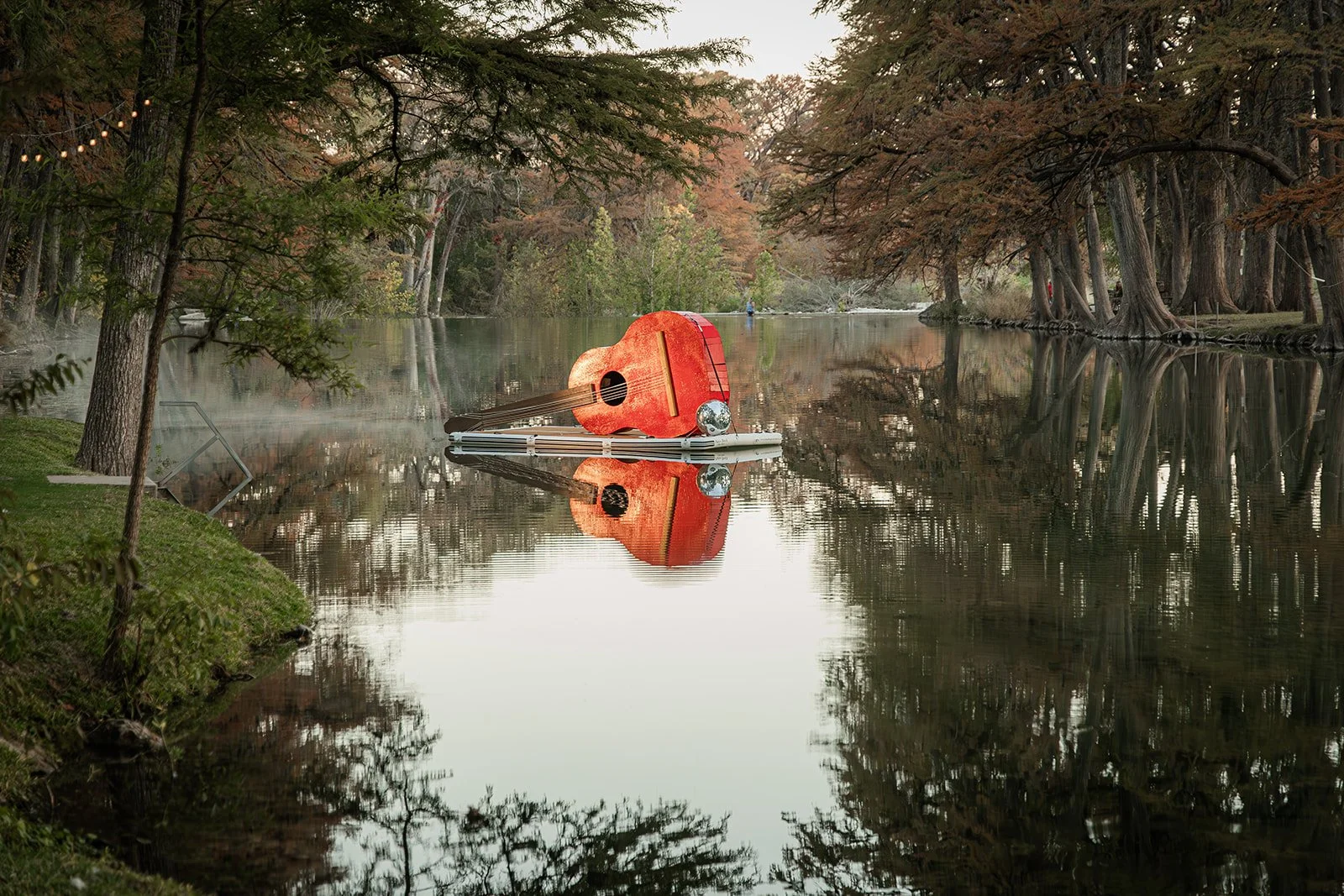 A lake surrounded by trees with a fallen acoustic guitar on a floating dock, reflecting on the water, and some mist rising from the lake.