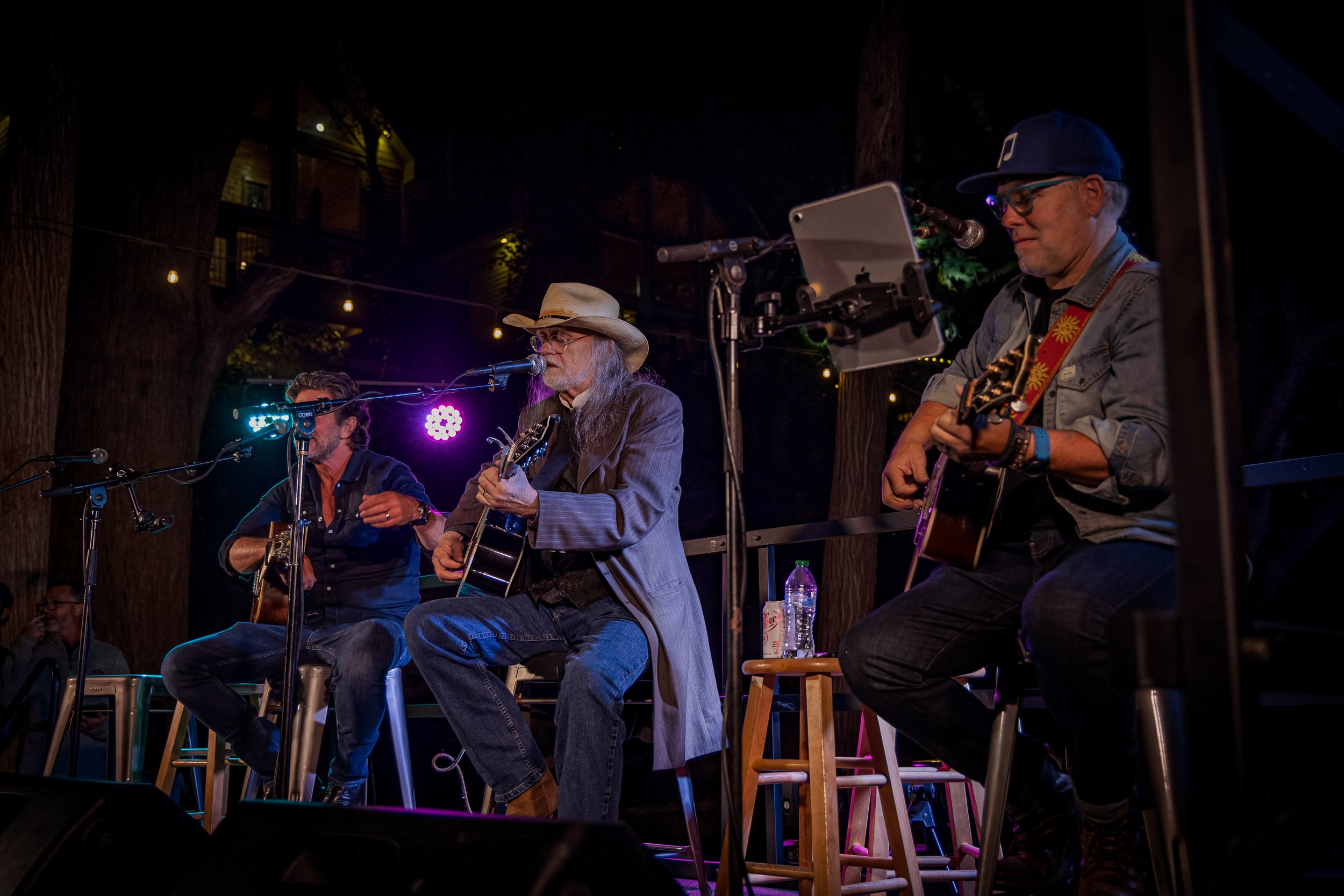 Three musicians performing on an outdoor stage at night, one playing guitar and singing into a microphone, wearing a cowboy hat; another playing guitar and wearing sunglasses and a cap; the third also playing guitar. String lights are hanging in the