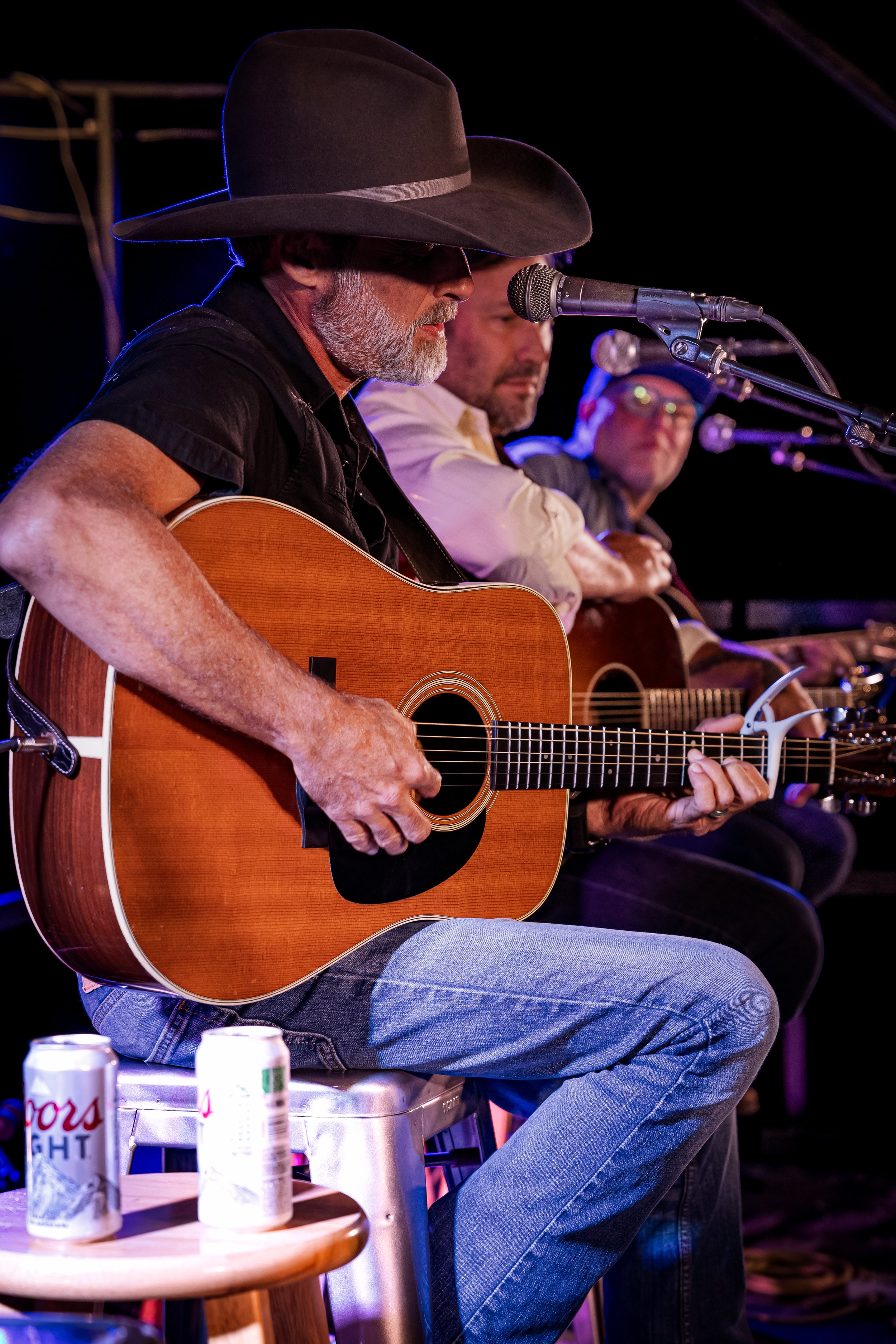 Three men playing guitars and singing into microphones on stage, with cans of Coors Light beer on a small table in front of them.