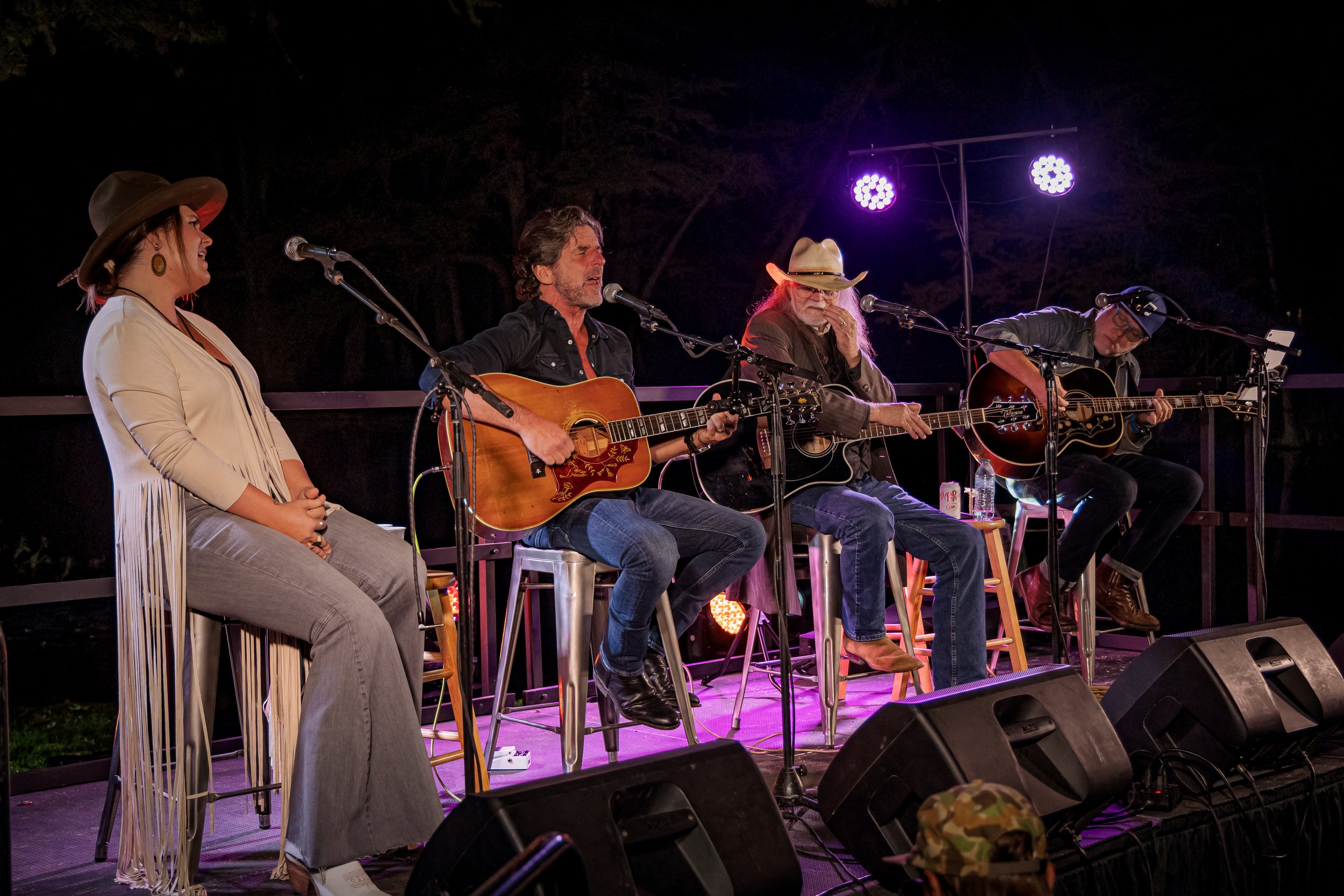Four musicians performing on a stage at night, three playing guitars and one singing into a microphone, with stage lighting and audio equipment.