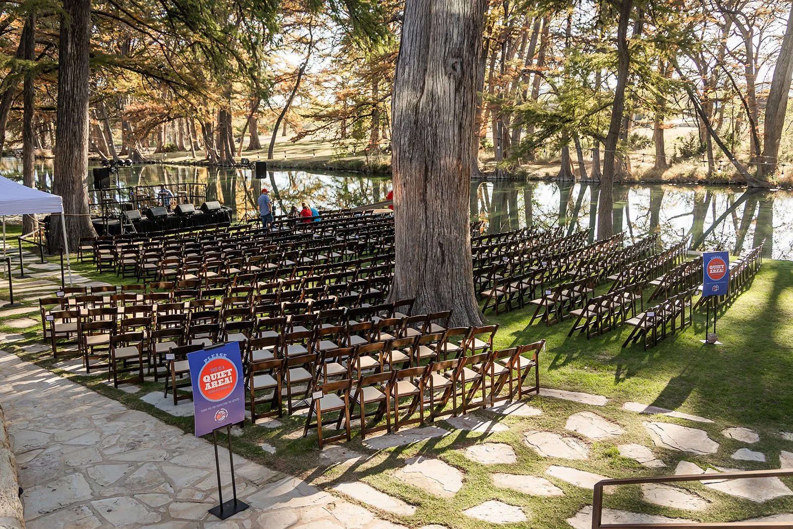 Outdoor venue setup by a water body with rows of chairs, a stage with sound equipment, and signs indicating a quiet area, surrounded by tall trees and reflecting water.