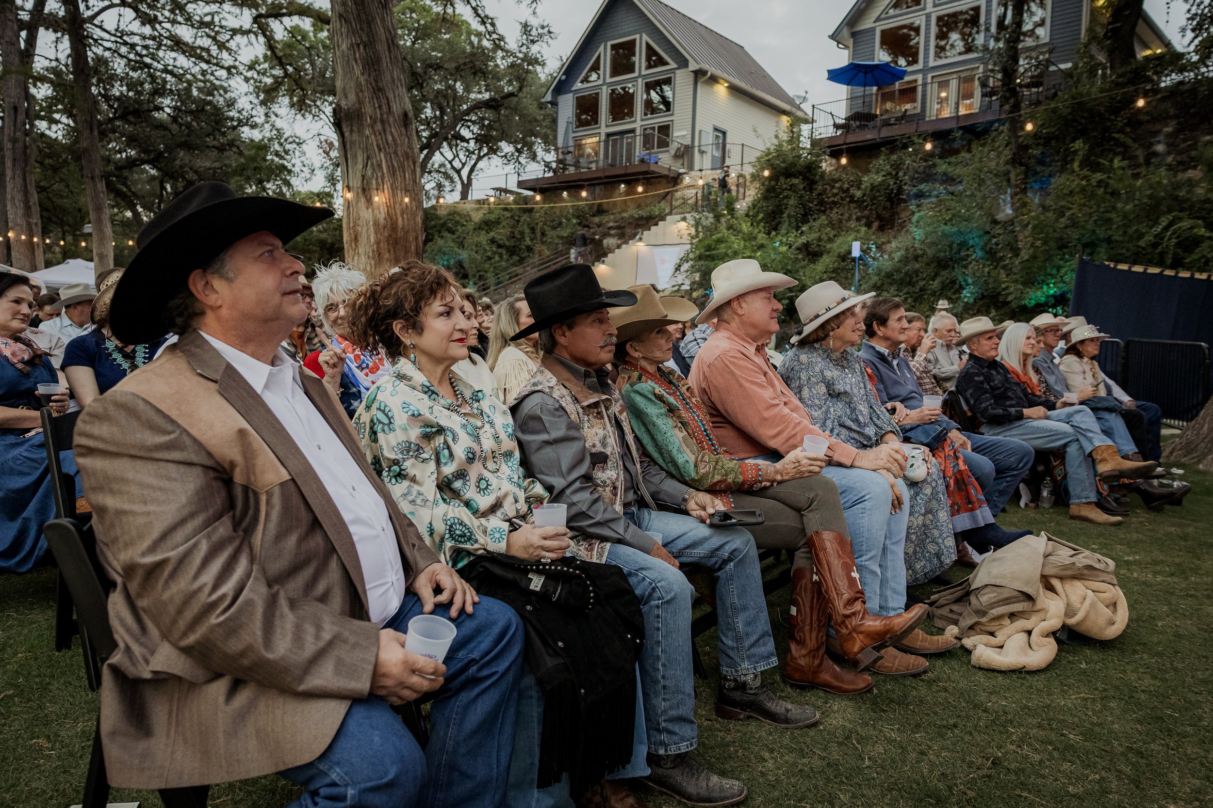 Audience of adults sitting outdoors, dressed in western-style attire such as cowboy hats, in front of a multi-story house with trees and string lights.