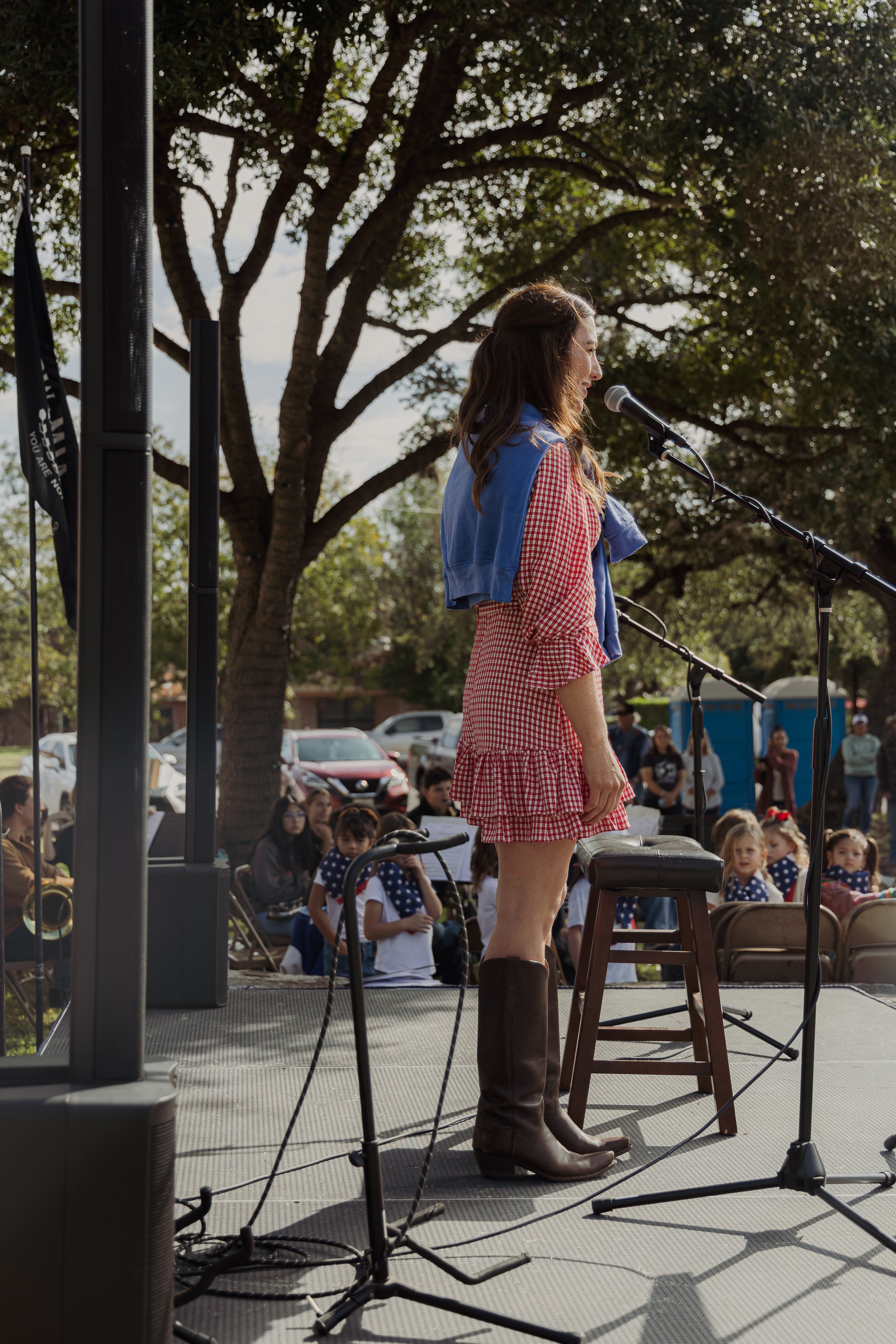Woman in red and white checkered dress, knee-high boots, and a blue shirt tied around shoulders, standing at microphone on outdoor stage, speaking to audience of children and adults under trees.