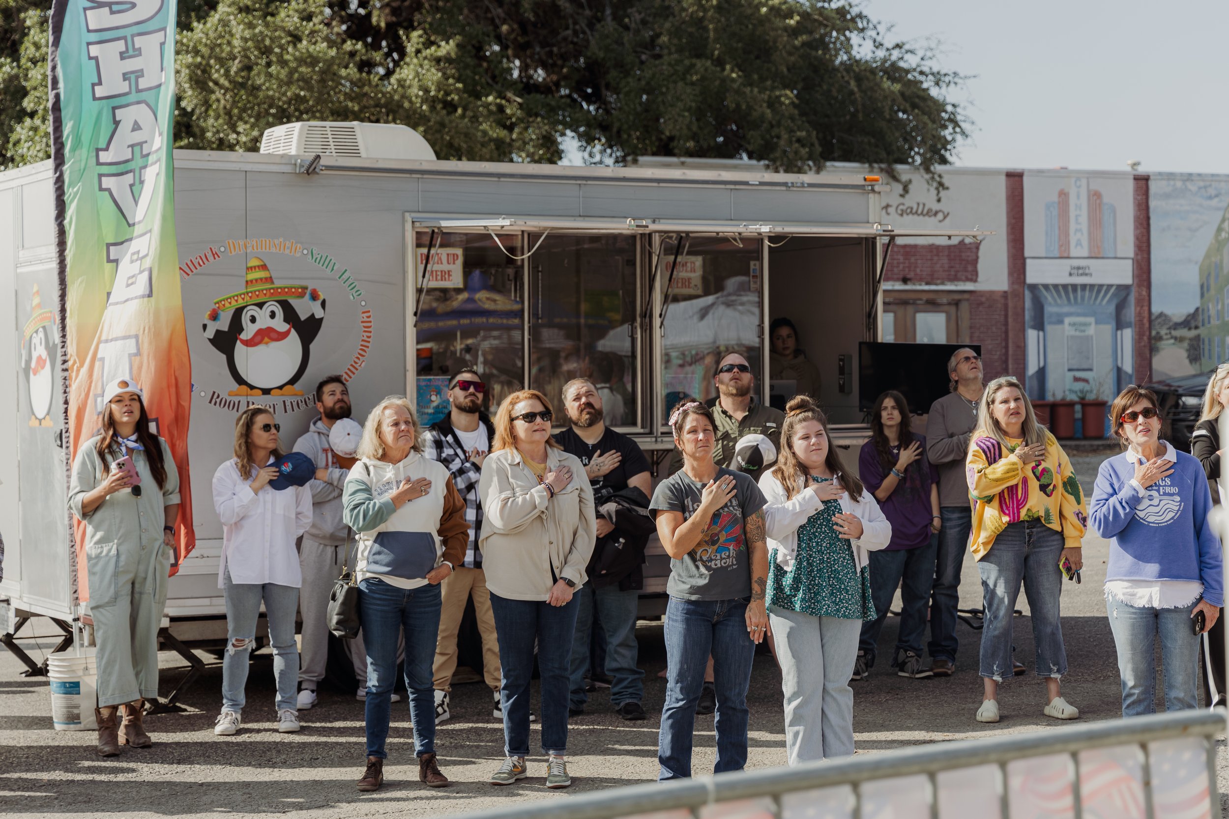 A group of people standing outdoors with their right hands over their hearts, participating in a patriotic or ceremonial event. Some are wearing casual clothing, and one person is dressed in a white coat. The background features a food truck with a c