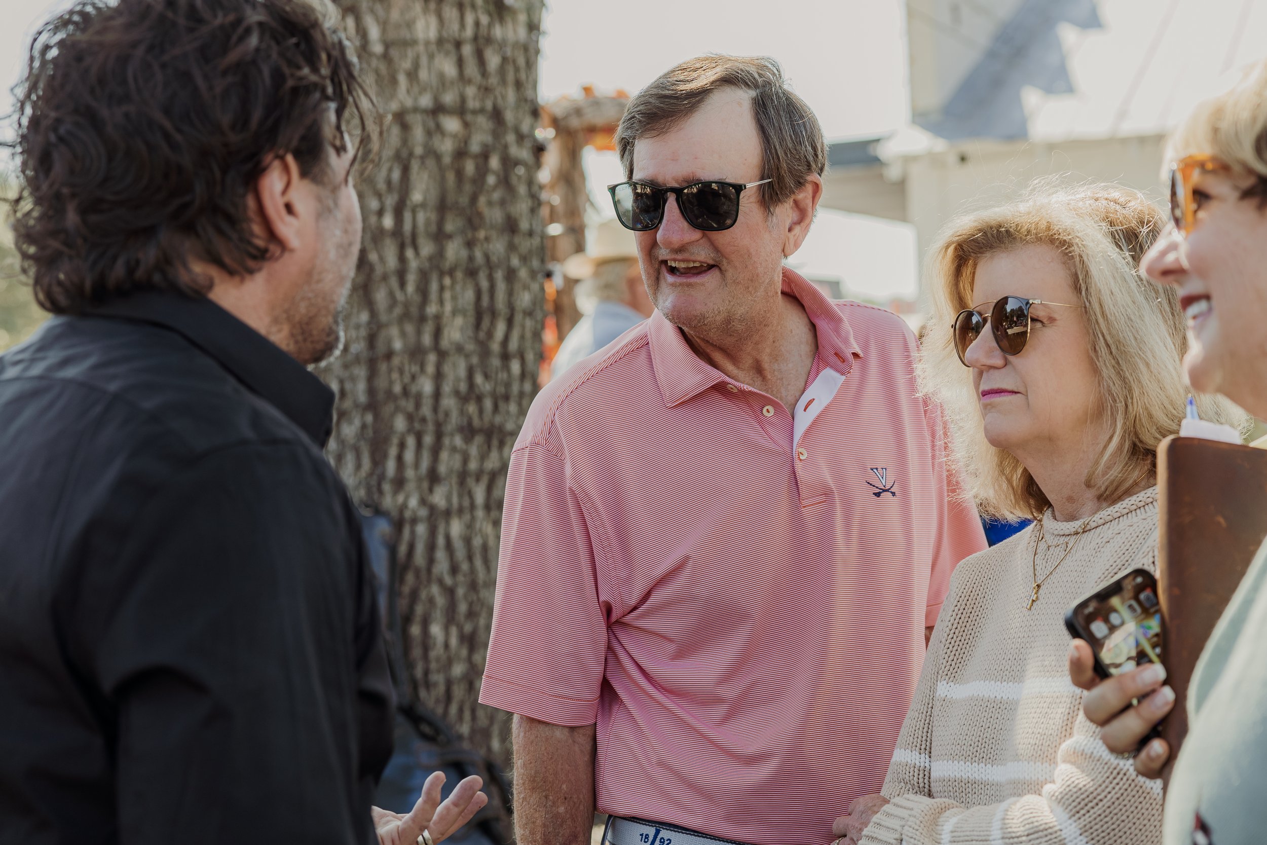 Group of four people talking outdoors, some wearing sunglasses, with trees and tents in the background.