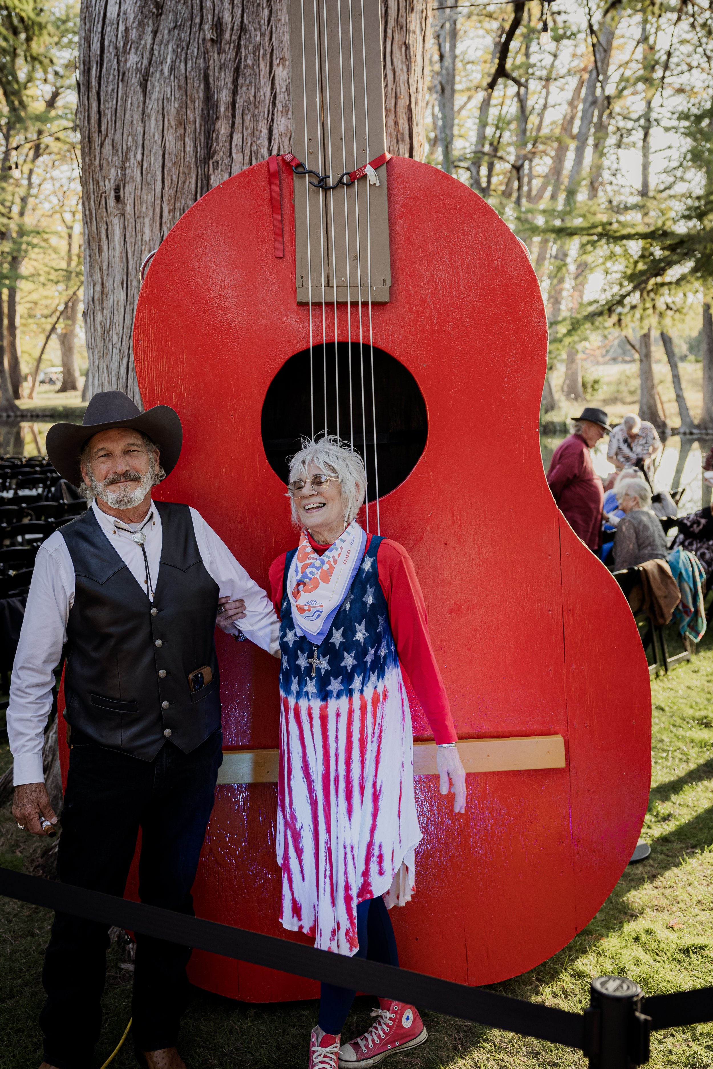 Two people standing in front of a large red guitar-shaped sculpture outdoors with trees and other people in the background.