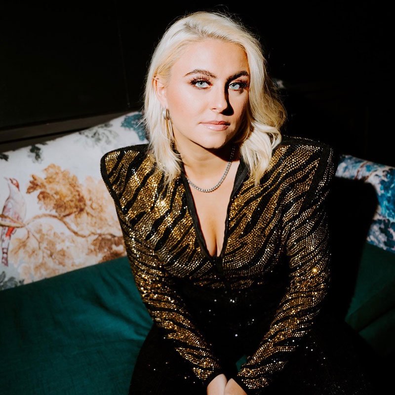 A woman with platinum blonde hair sitting on a green couch in a dimly lit room, wearing a black and gold sequined outfit and a silver necklace.