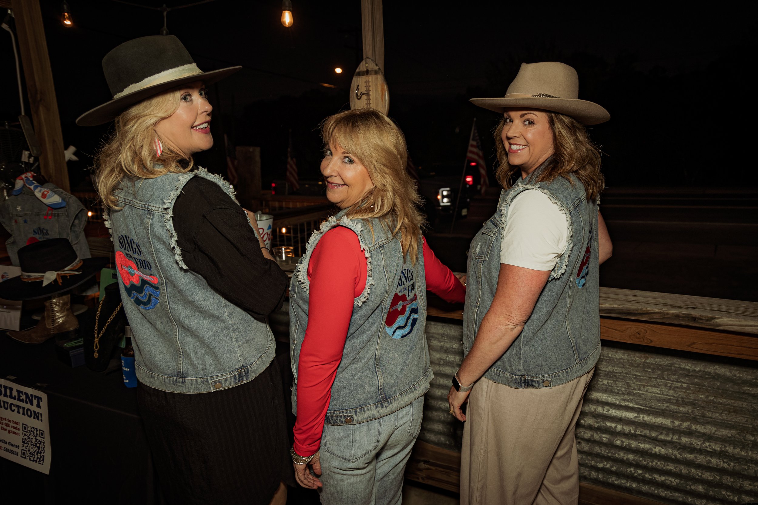 Three women wearing denim vests with Tros and guitar patches, cowboy hats, and casual outfits, standing together at a nighttime outdoor event.
