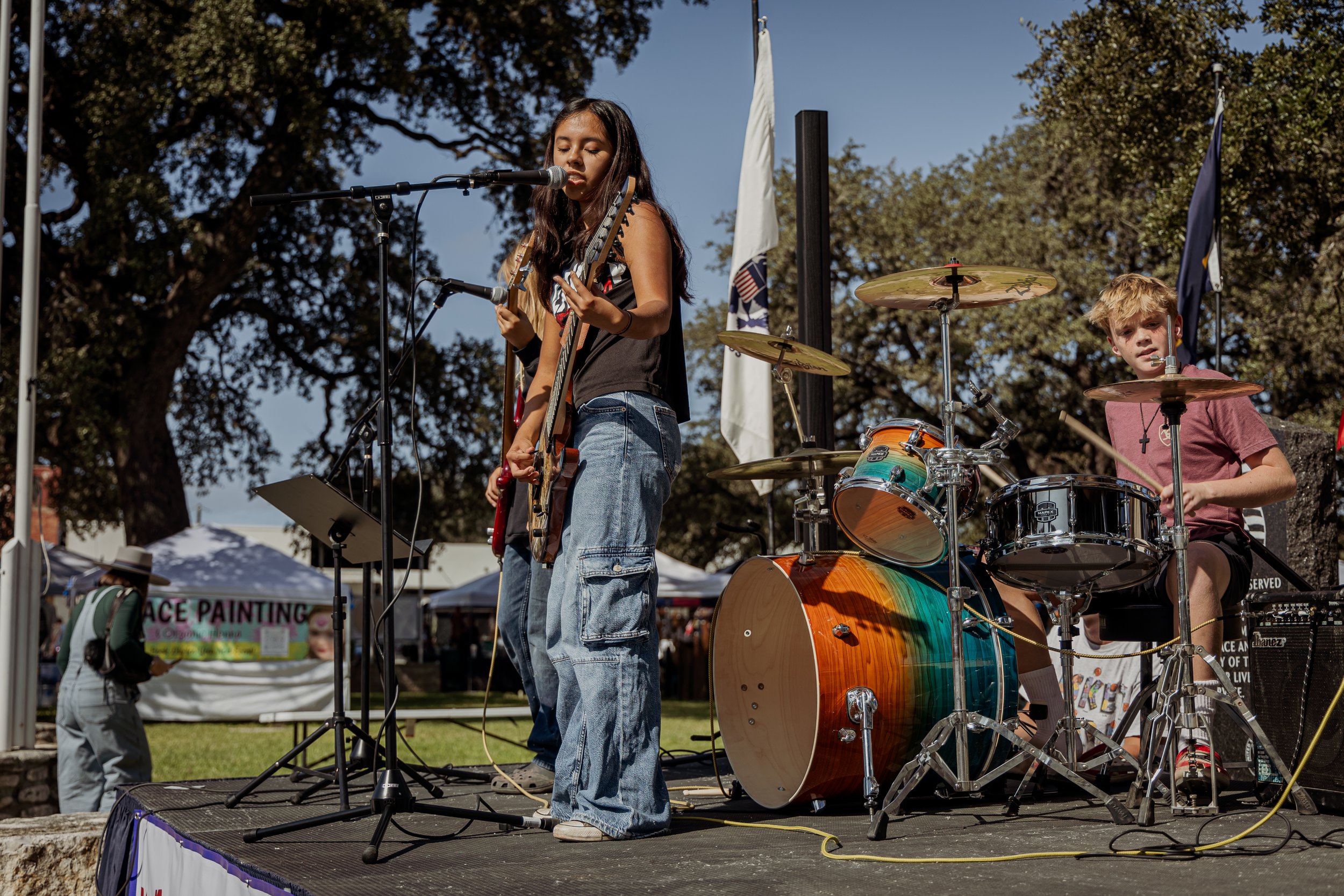 Two young musicians performing on an outdoor stage: a girl singing and playing an electric guitar, and a boy playing a drum set, with tents and trees in the background.