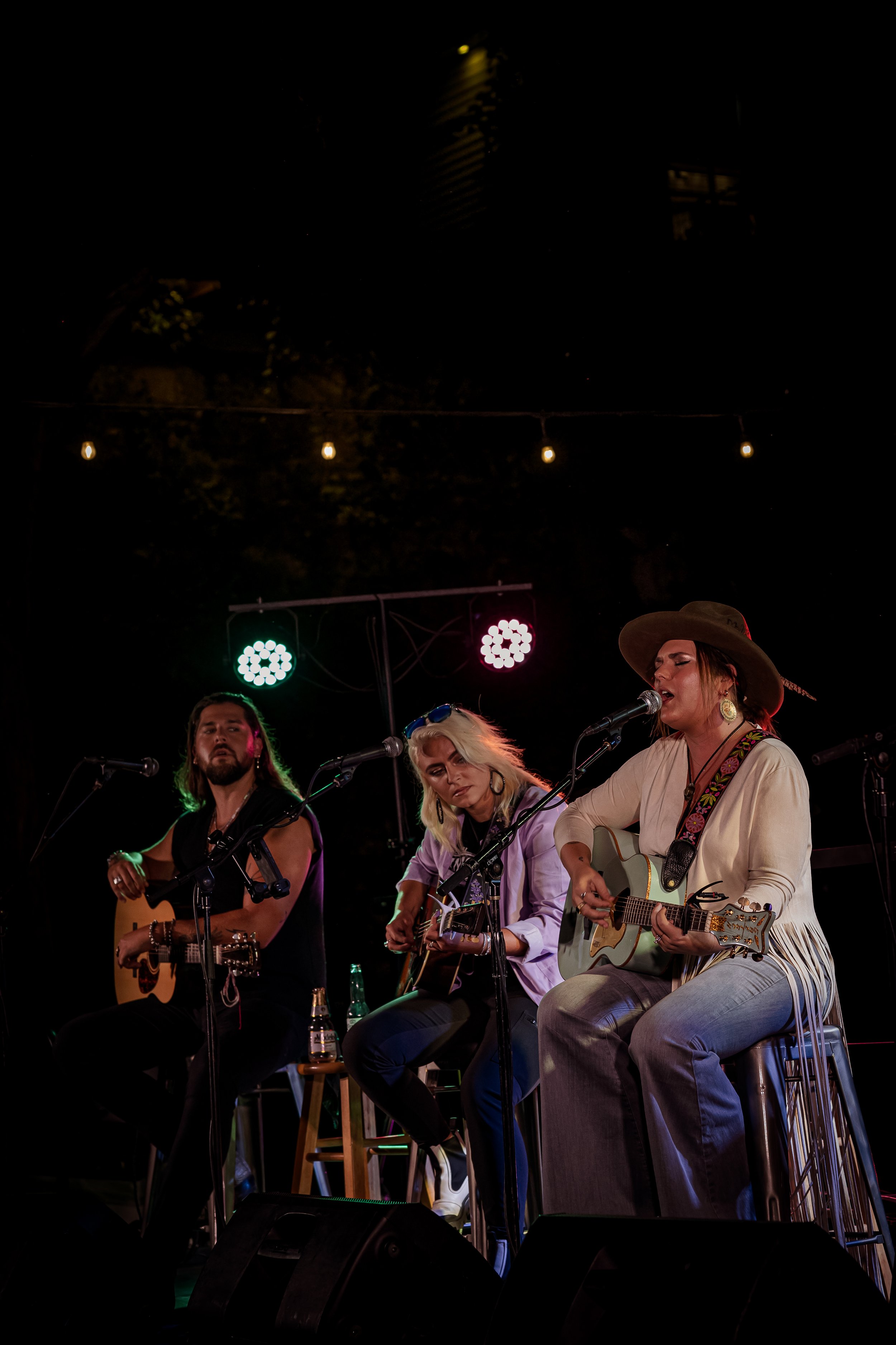 Three women play acoustic guitars on stage, singing into microphones during a live outdoor music performance at night, with colorful stage lights and string lights overhead.