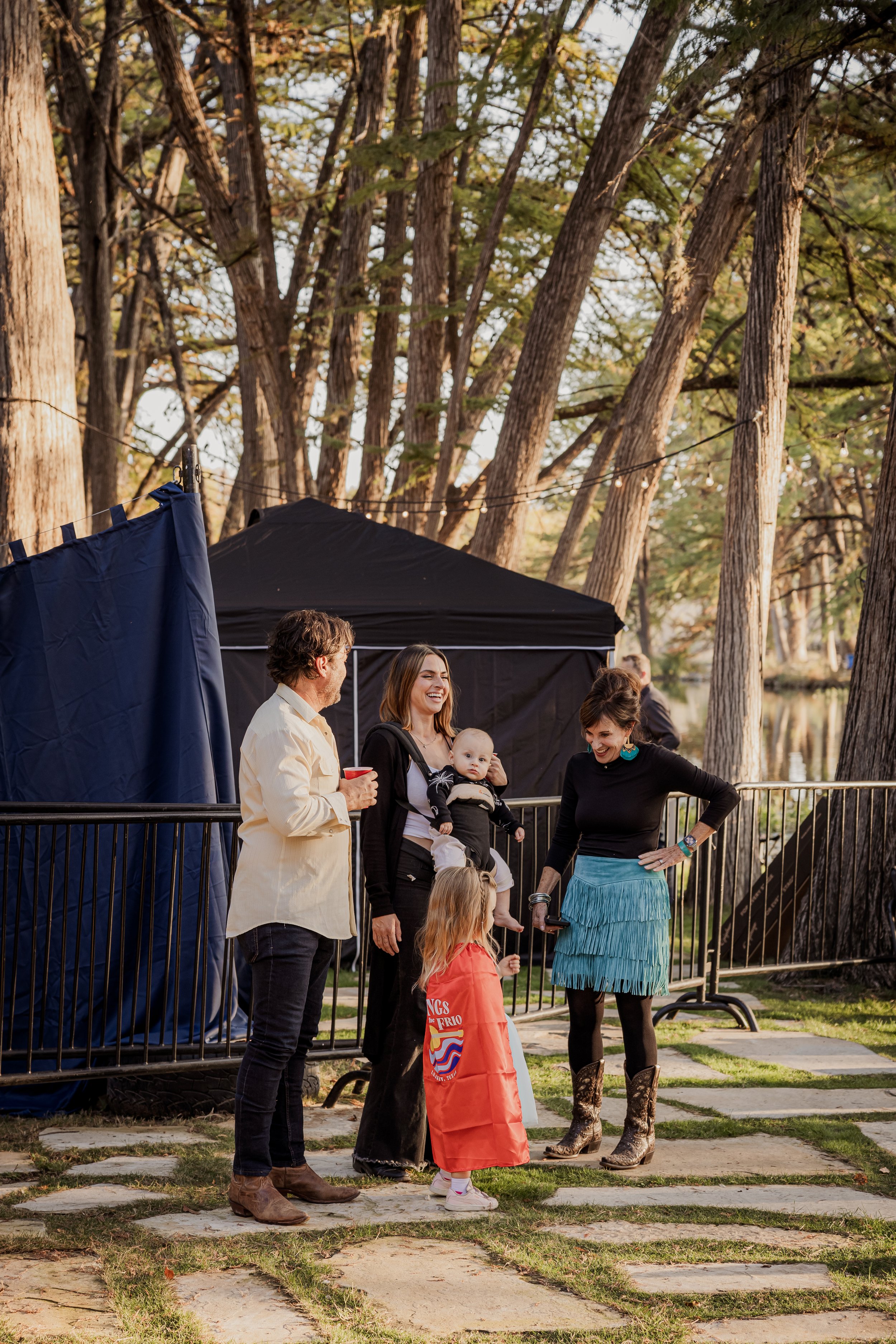 Group of five people, including children, chatting outdoors near a black tent and a railing, surrounded by tall trees during autumn.