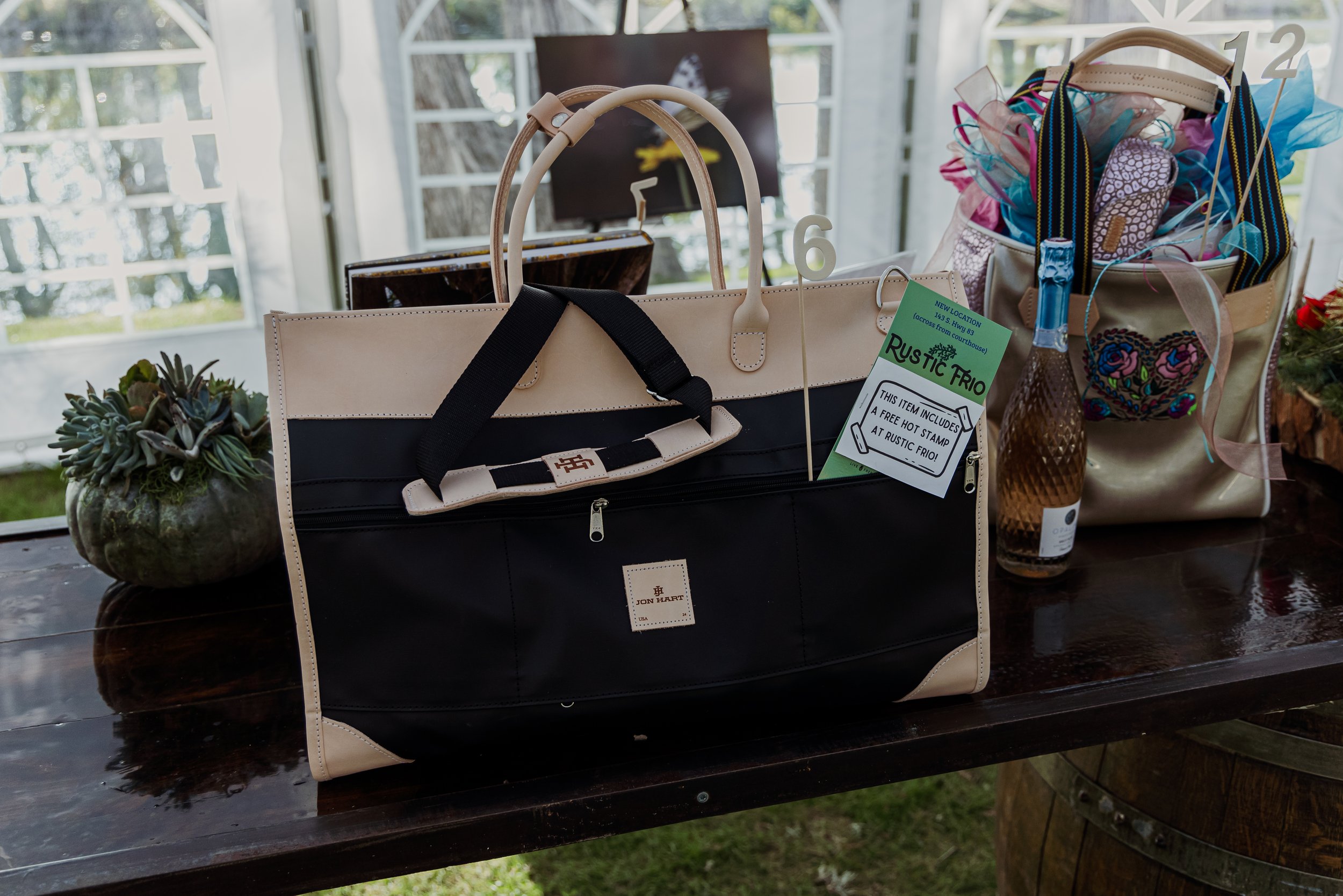 A black and beige Jon Hart tote bag on a wooden table, surrounded by potted succulent and decorative bags, with a bottle and a raffle ticket attached.