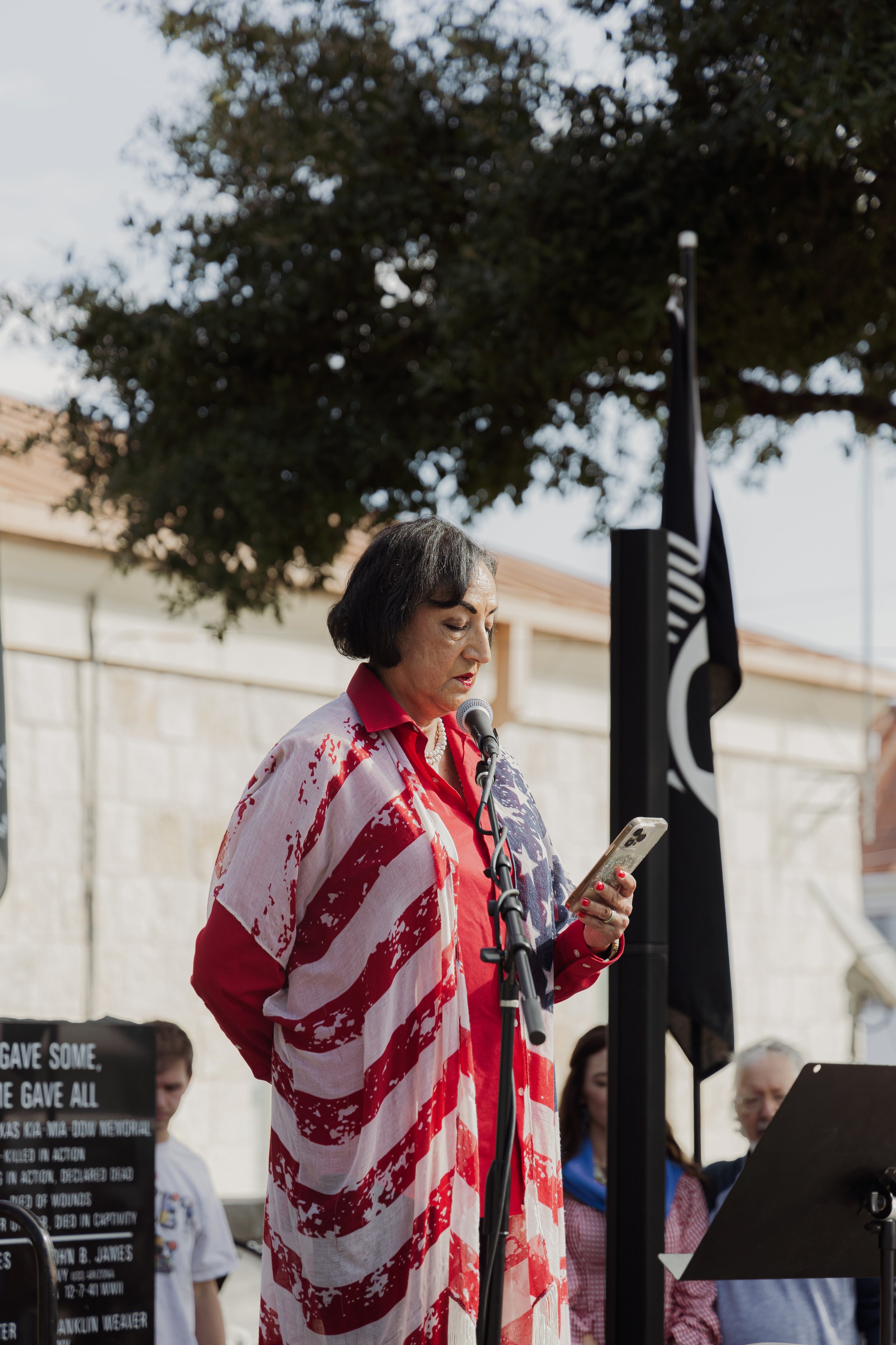 A woman standing at a microphone during a public outdoor event, wearing a red shirt and an American flag patterned shawl, with people and a flag in the background.