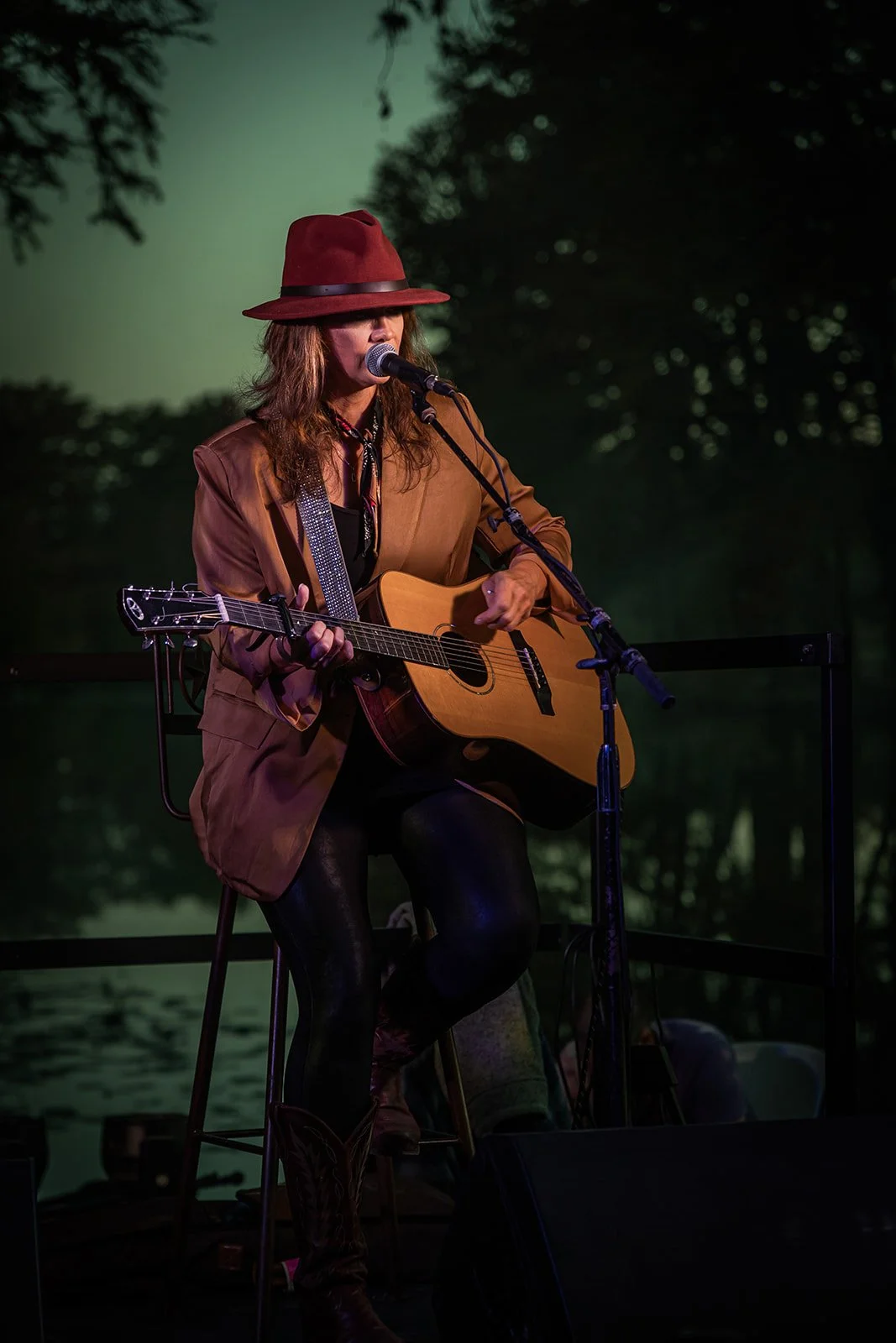 A female musician performing outdoors at night, sitting on a stool, playing an acoustic guitar and singing into a microphone, wearing a brown hat, a brown jacket, and black pants, with trees and a water body in the background.