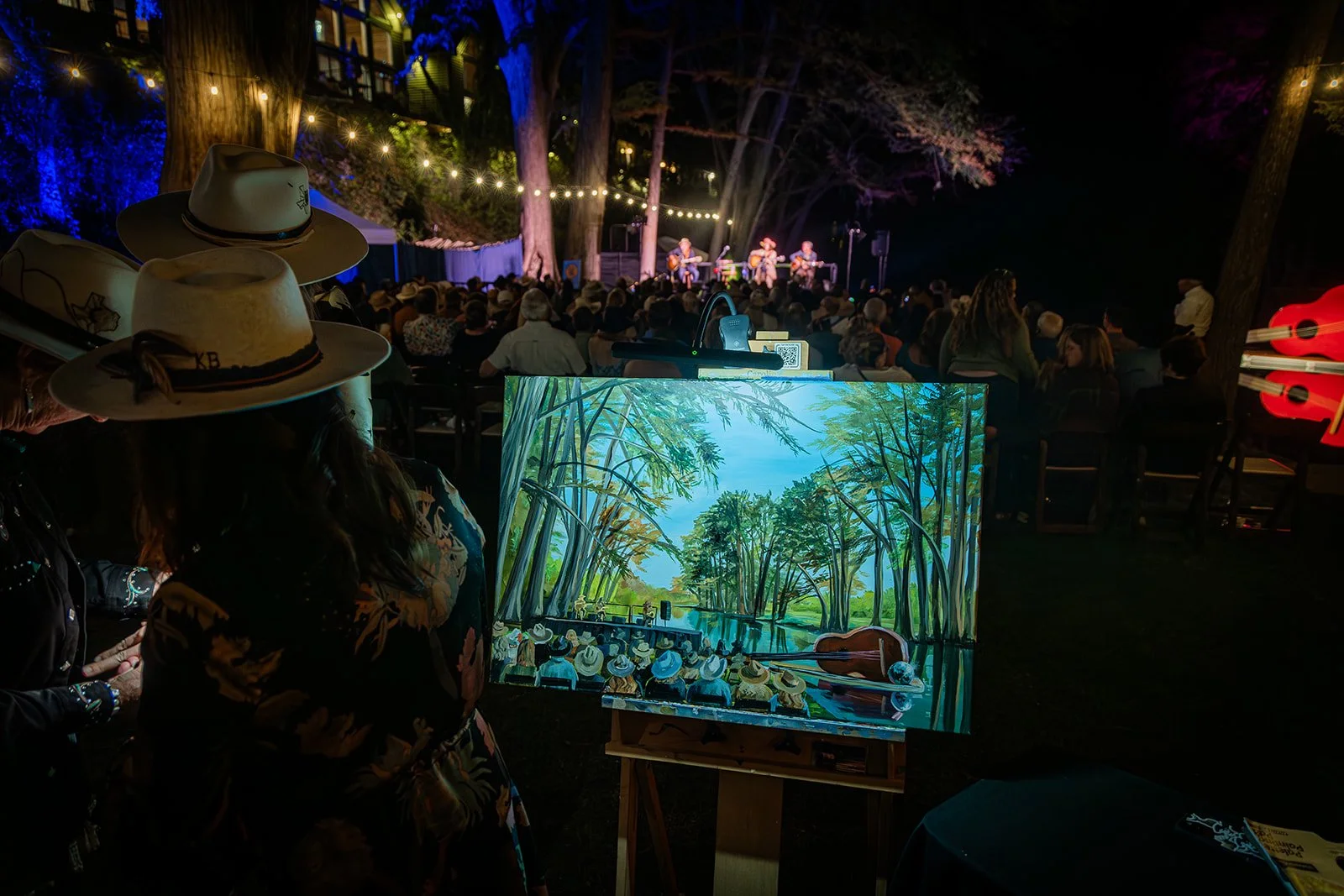 An outdoor concert at night with a crowd watching a band perform on stage, string lights hanging overhead, and a painting of a natural scene of trees and water prominently displayed in the foreground.
