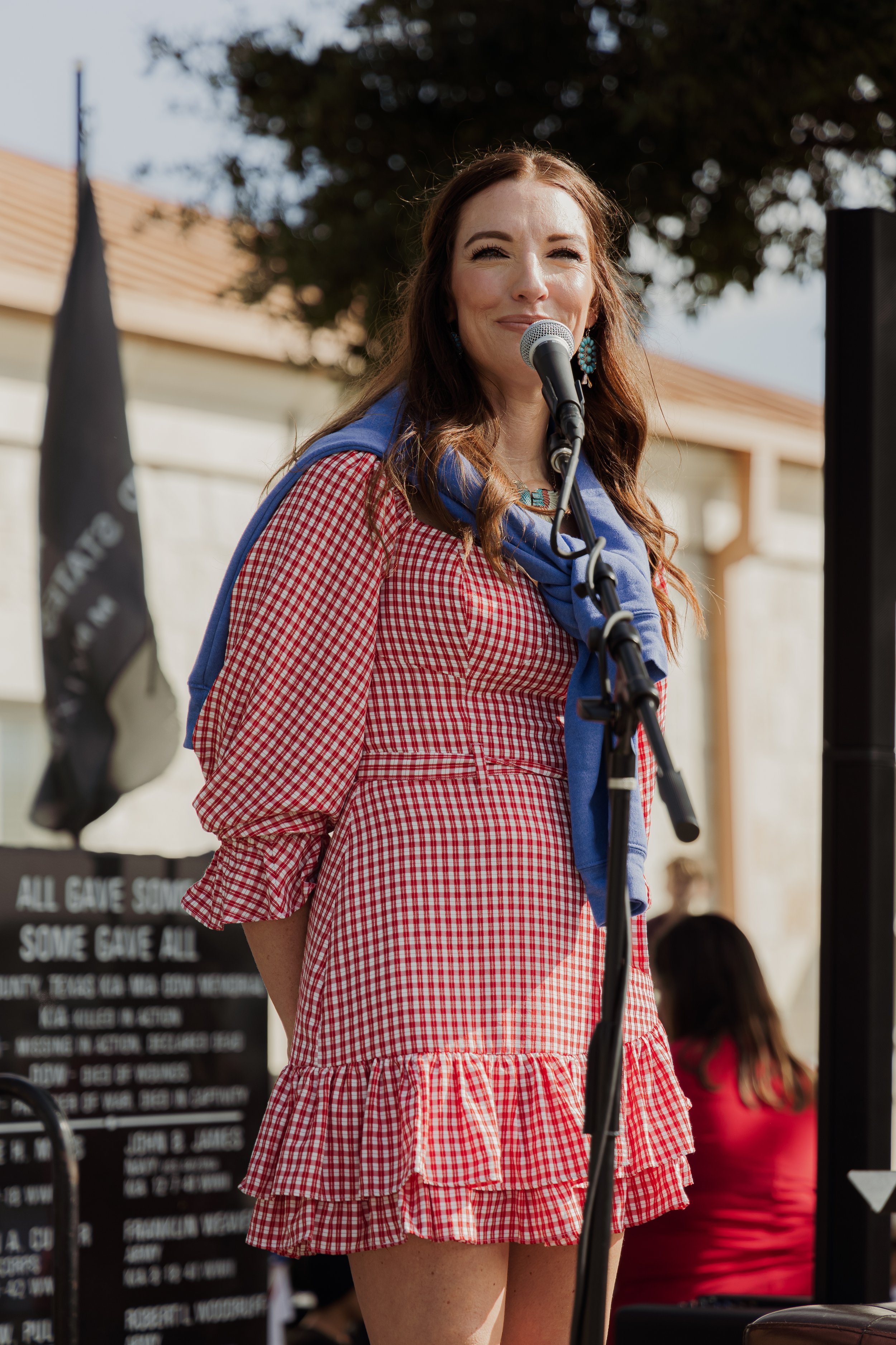 A woman in a red and white checkered dress with ruffled sleeves and hem, standing at a microphone outdoors, smiling.