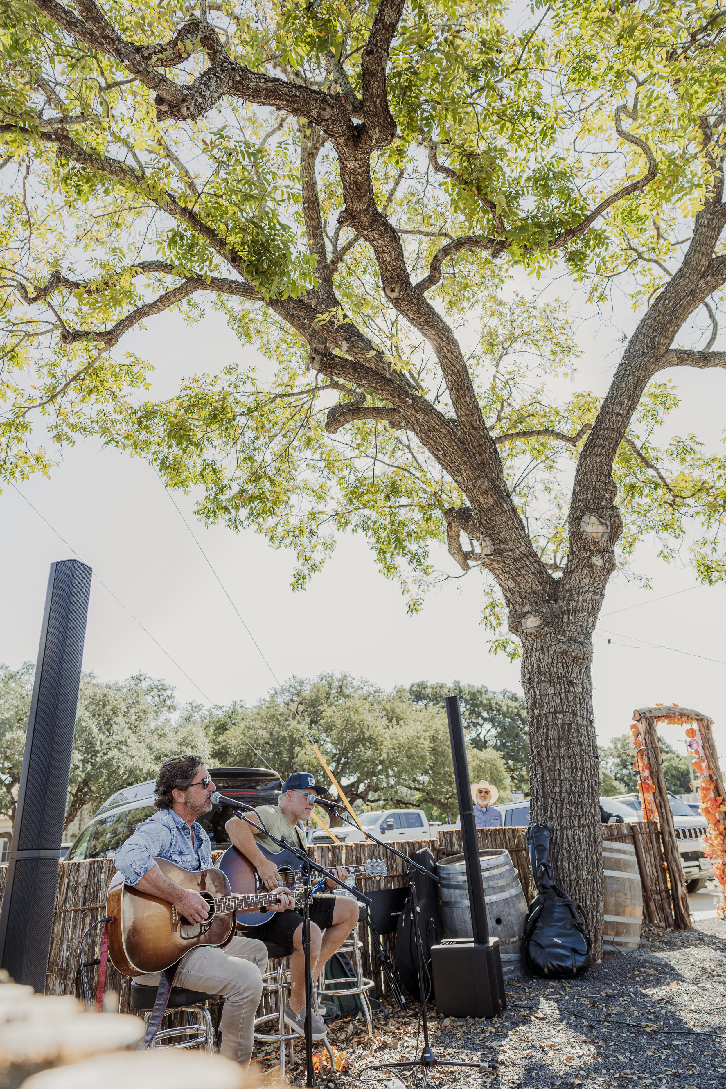 Two musicians playing acoustic guitars and singing outdoors under a large leafy tree, with a speaker and sound equipment nearby, and a person in the background wearing a hat.