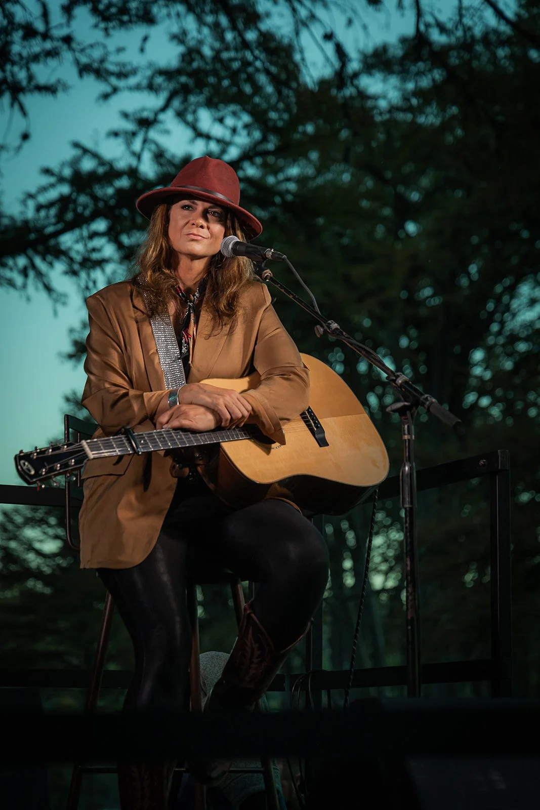 Woman sitting outdoors on a stage, holding an acoustic guitar, wearing a brown jacket and a red hat, with trees in the background.