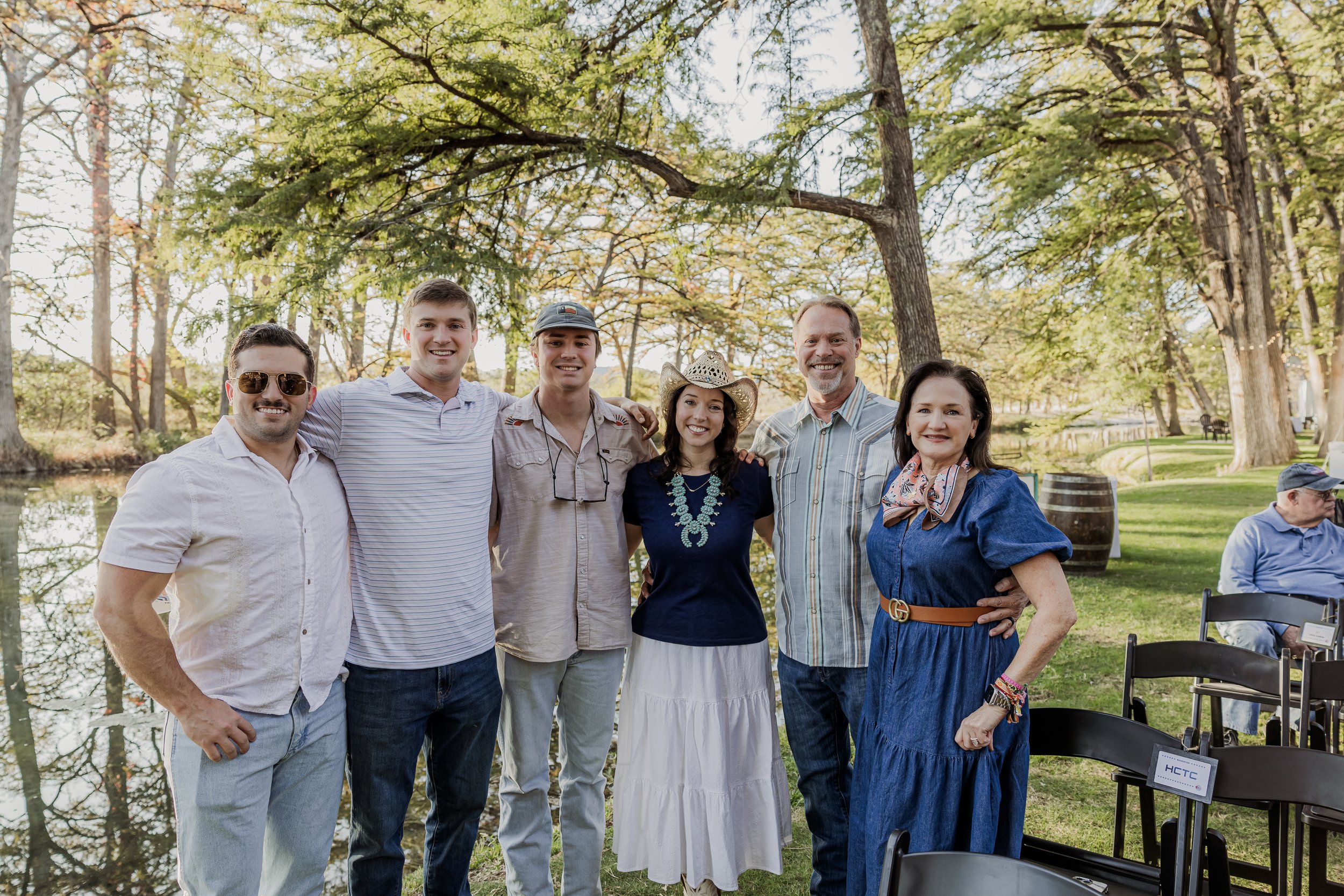 Group of seven people standing outdoors near a pond, smiling, in a wooded park during daytime.