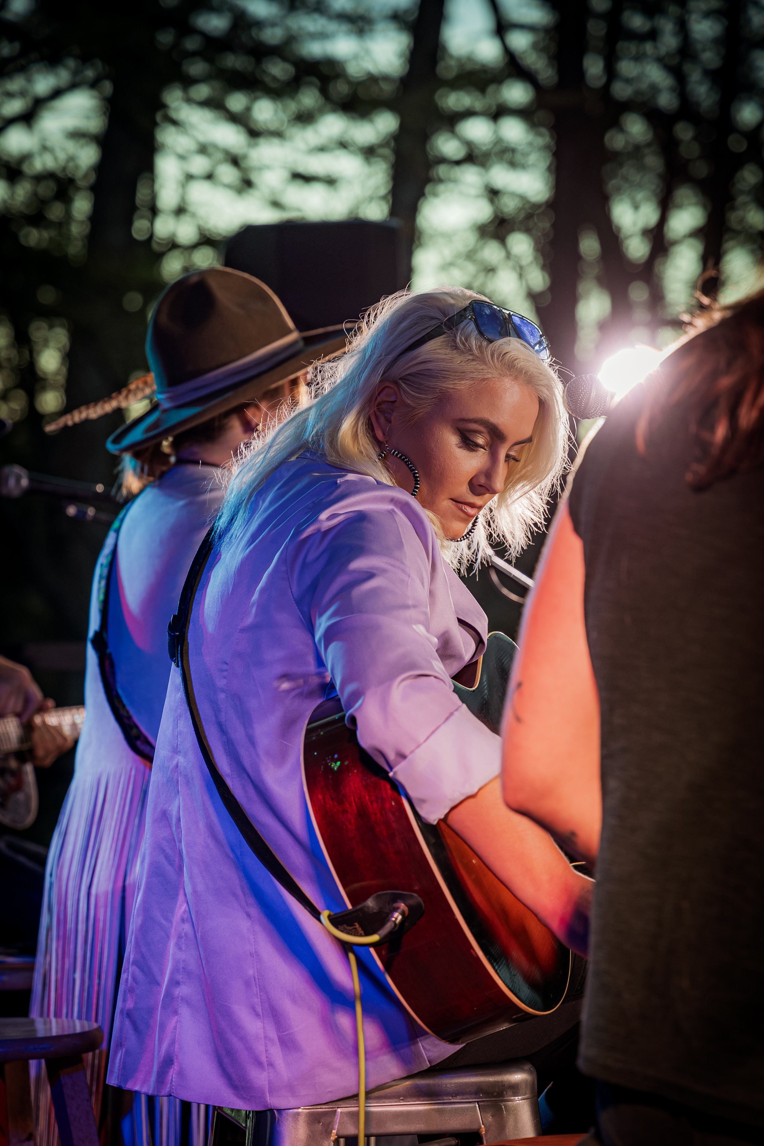 A woman playing an acoustic guitar on stage, surrounded by other musicians, during a live outdoor performance with trees in the background.
