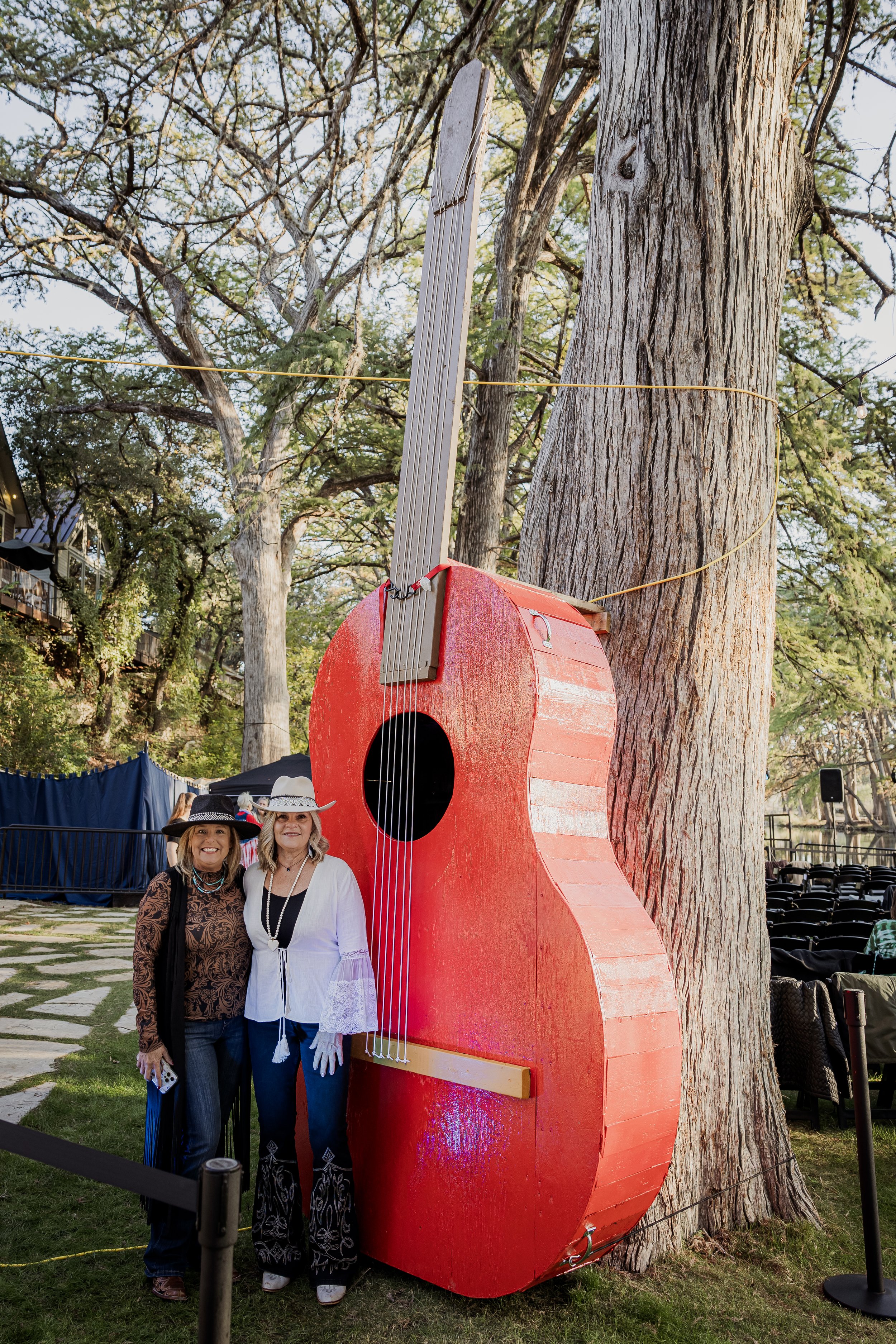 Two women dressed in cowboy hats and western attire standing beside a large red guitar sculpture outdoors near trees.