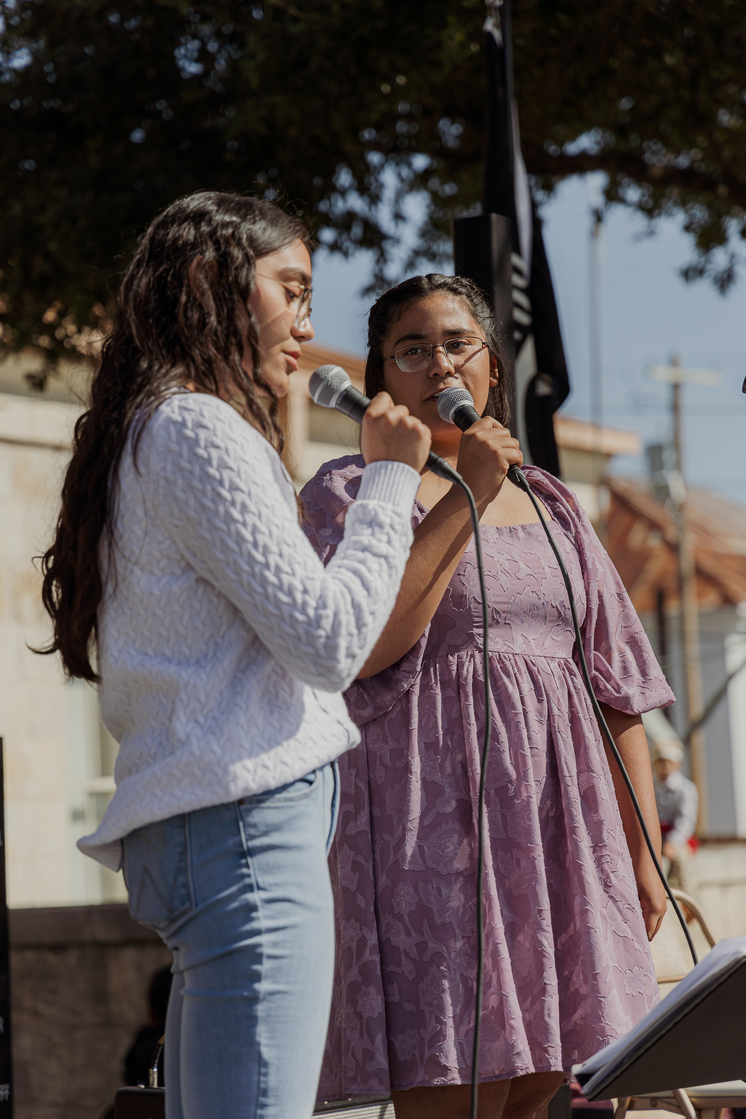 Two women speaking into microphones at an outdoor event, with a black flag and trees in the background.