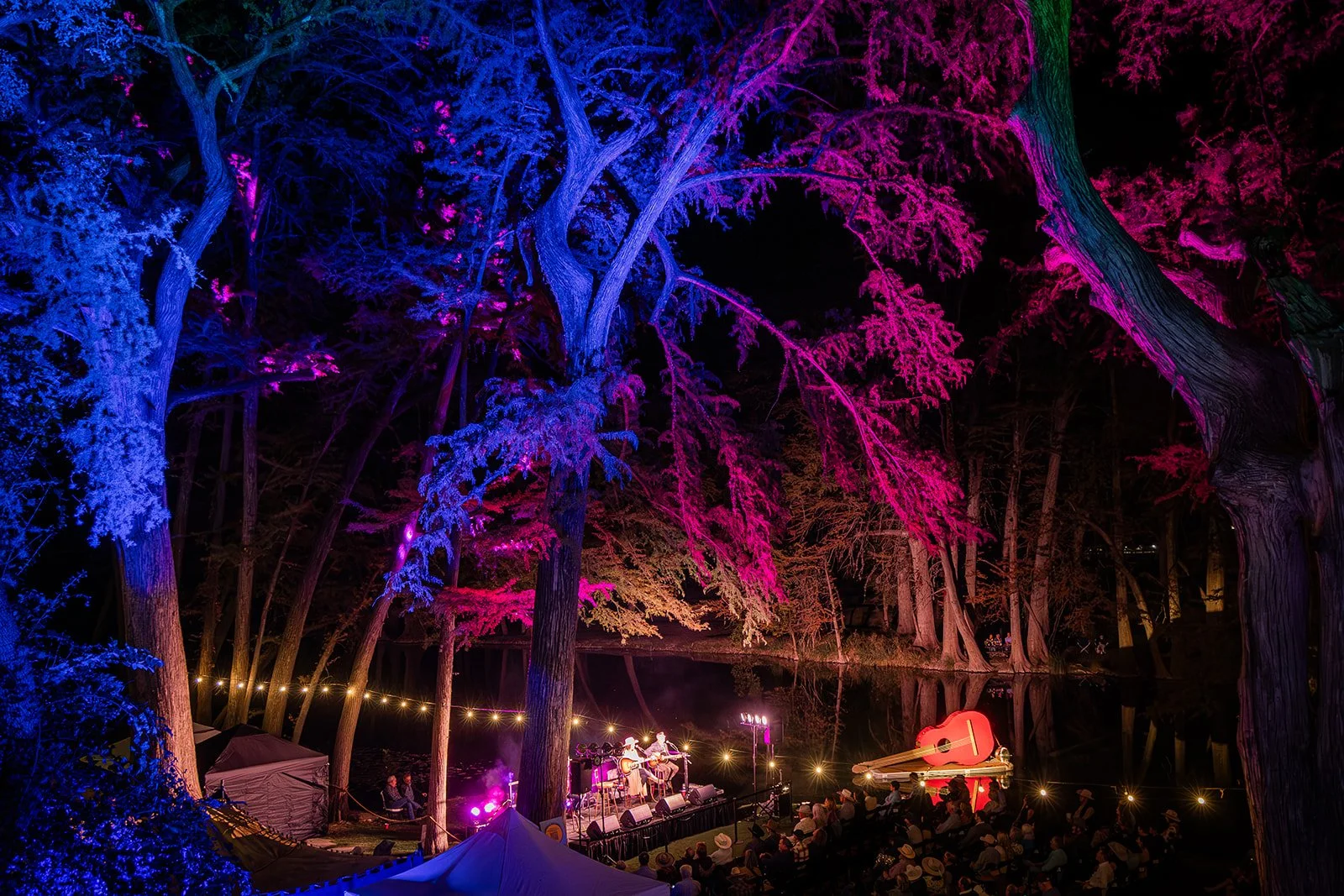 Nighttime outdoor concert by a pond, illuminated with colorful lights on trees, featuring a stage with musicians, an oversized guitar sculpture, and audience watching.